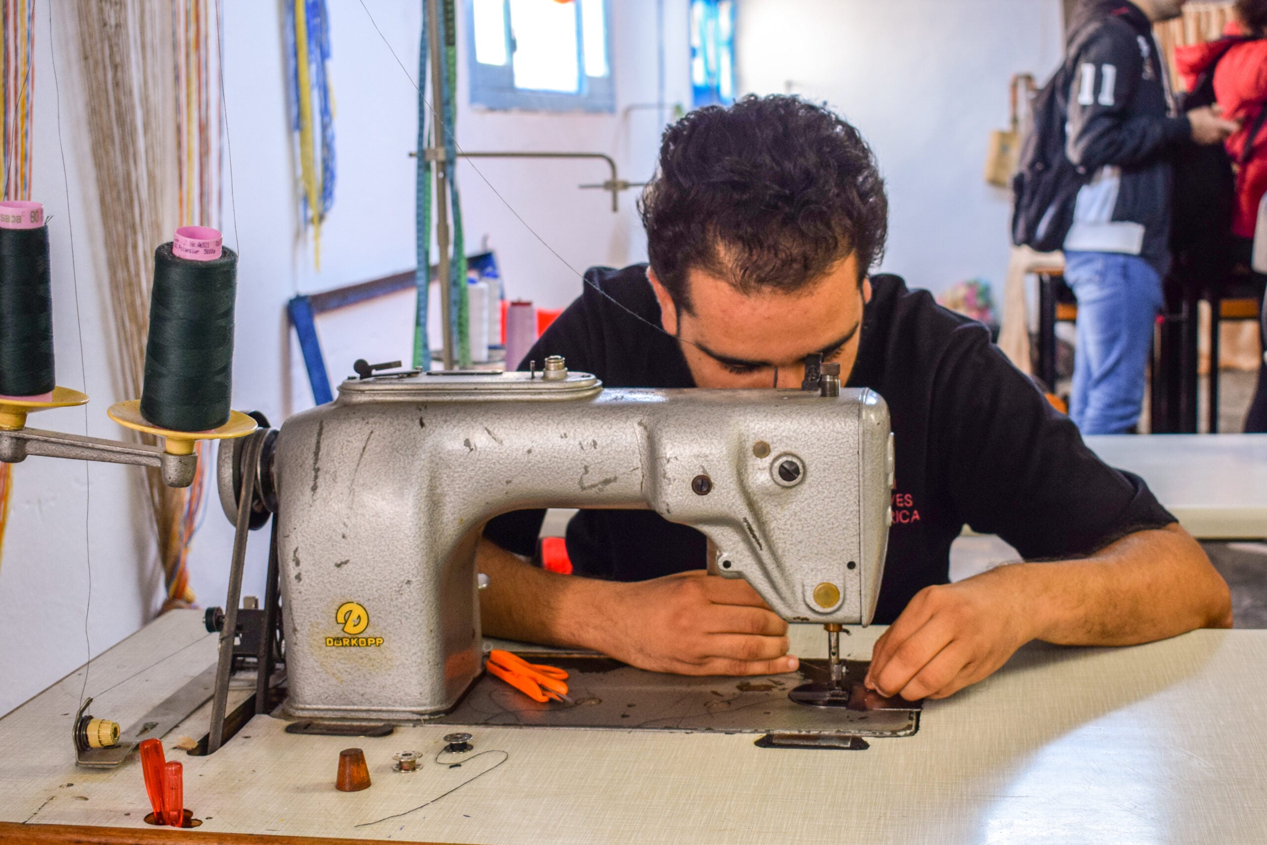 A person sits at a table, focused on threading or adjusting a sewing machine. Various sewing supplies and thread spools are visible nearby, and other people are blurred in the background.
