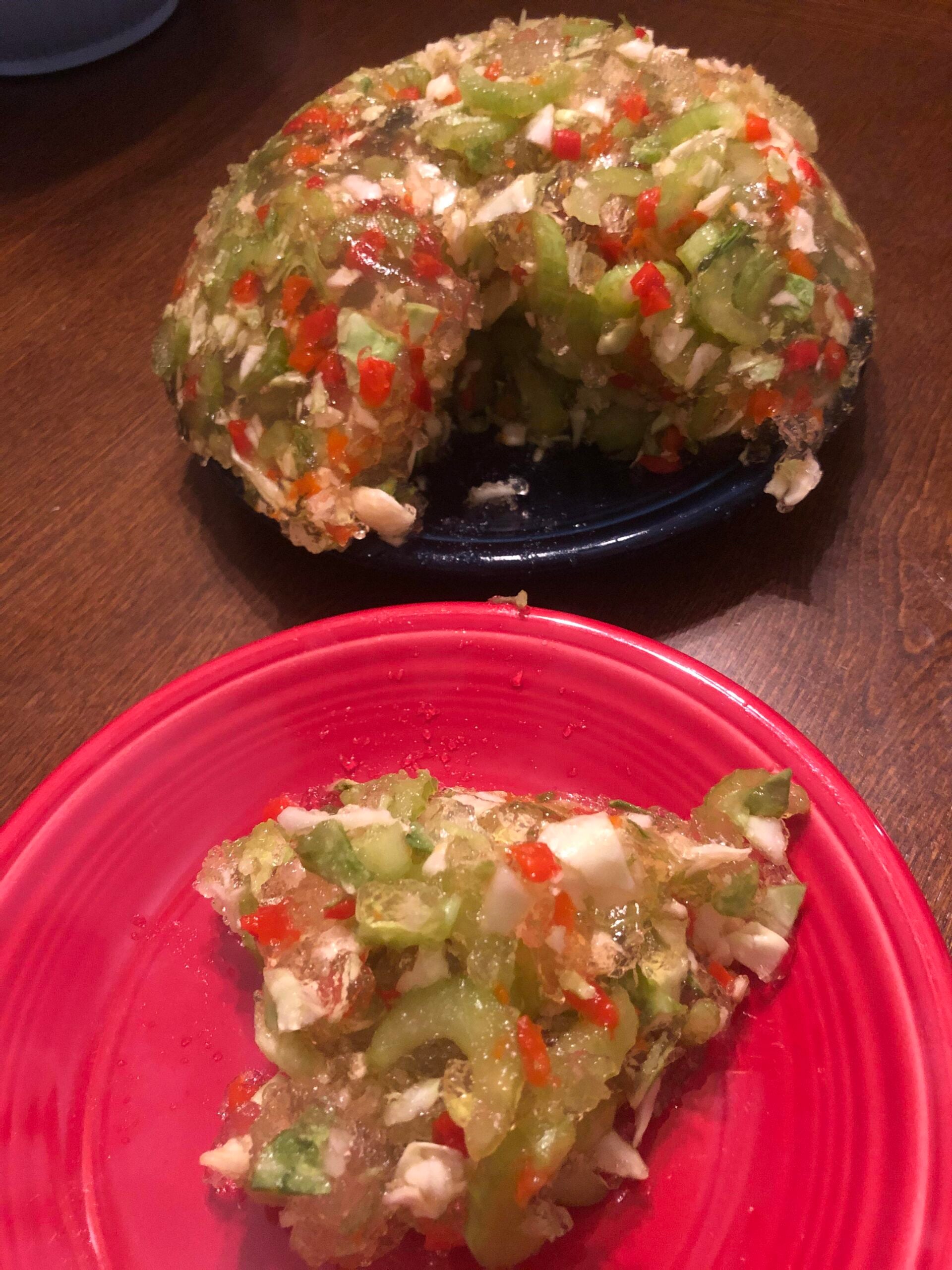 A gelatin salad with chopped vegetables and possibly eggs is shown, molded in a ring shape on a blue plate. A slice is served on a red plate in the foreground.