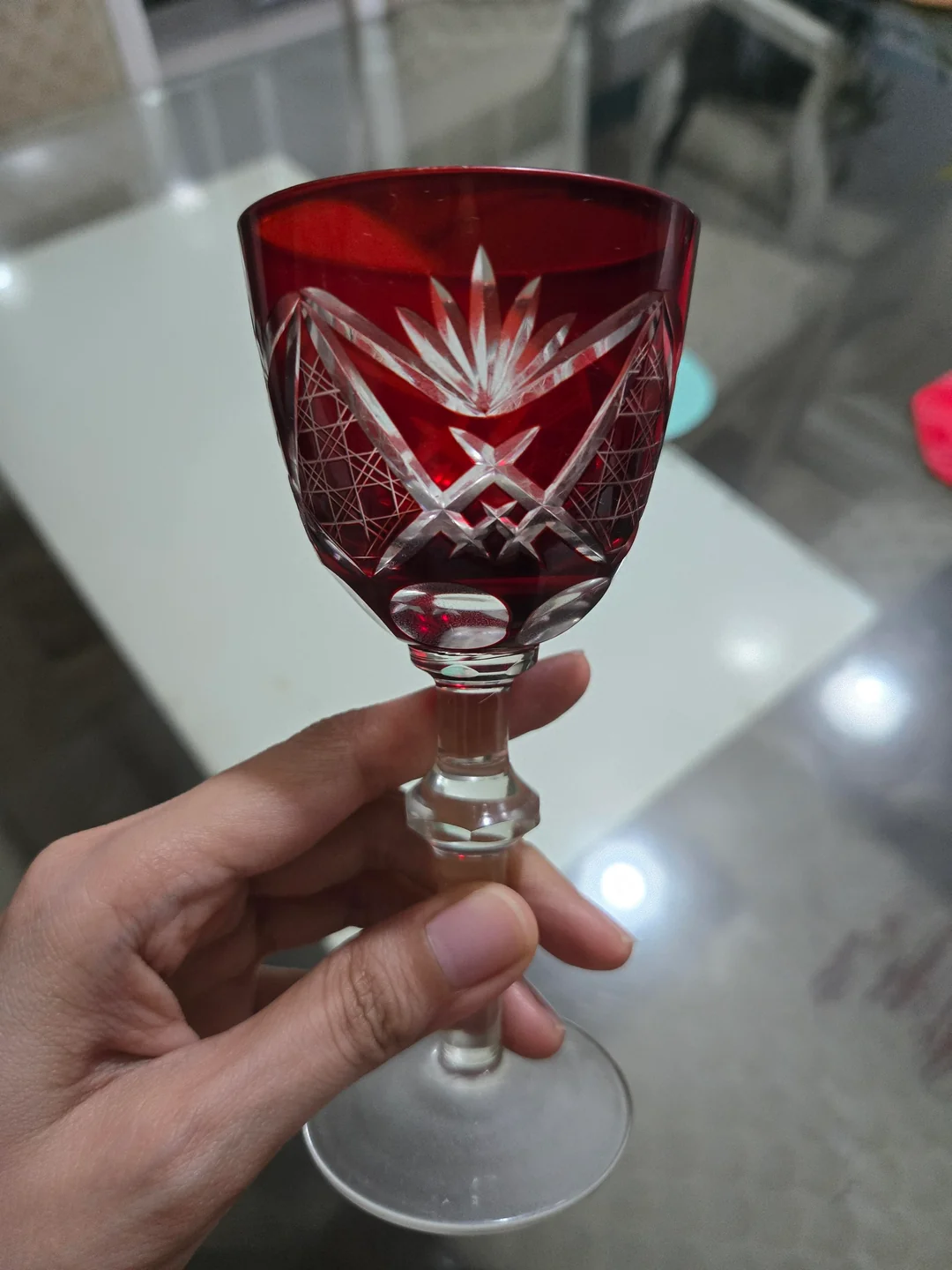 A hand holding a red and clear cut glass goblet with intricate geometric and starburst patterns, above a glass table.