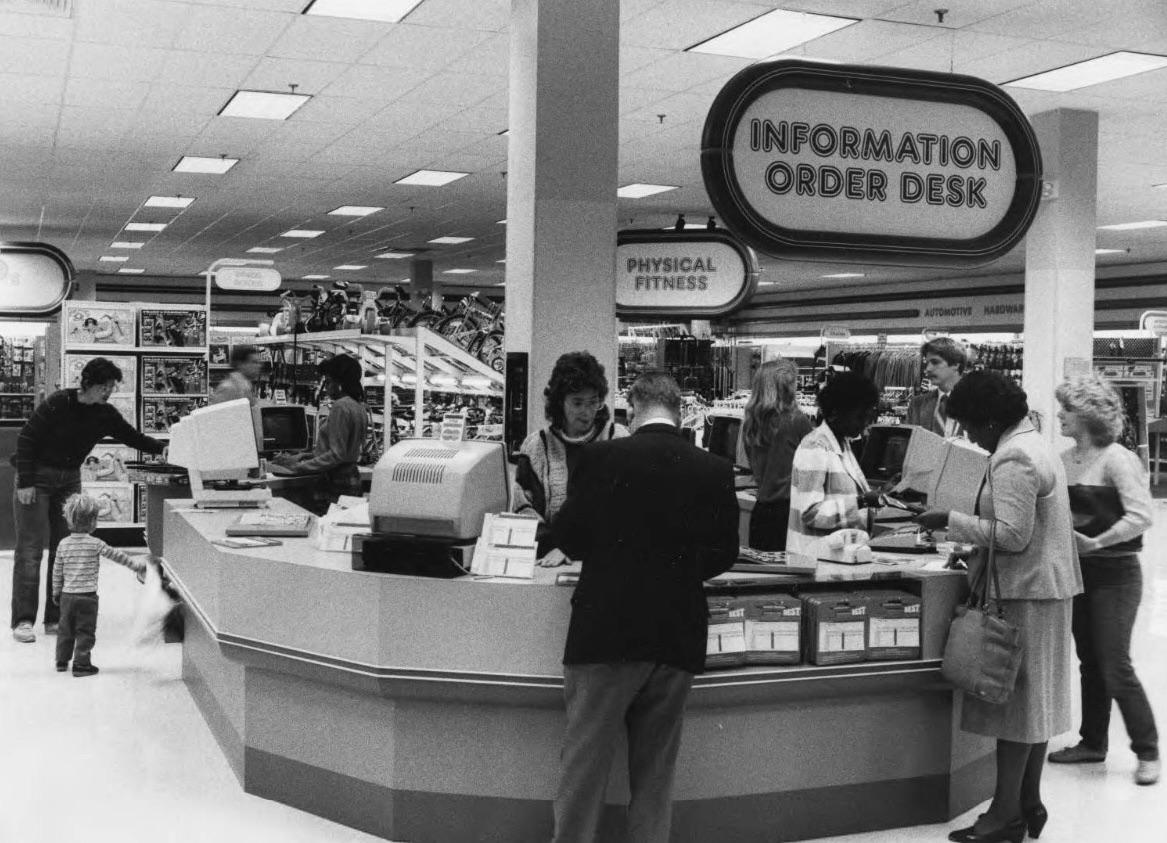 Black and white photo of people at an “Information Order Desk” in a retail store. Several customers speak with staff behind the counter; shelves and various products are visible in the background.