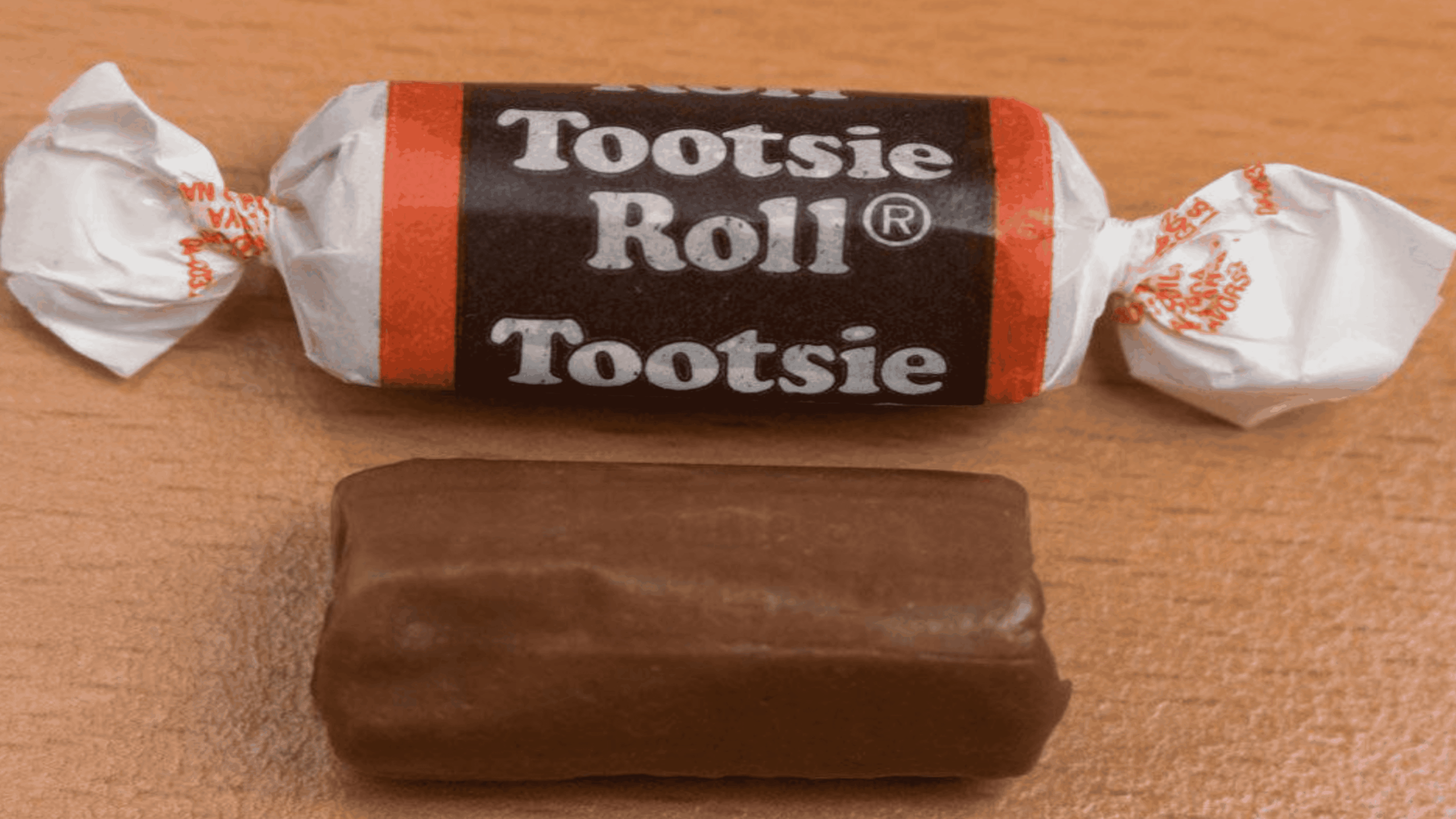 Unwrapped and wrapped Tootsie Roll candies on a wooden surface; the unwrapped piece is rectangular and chocolate-colored, while the wrapped one has white and black packaging.