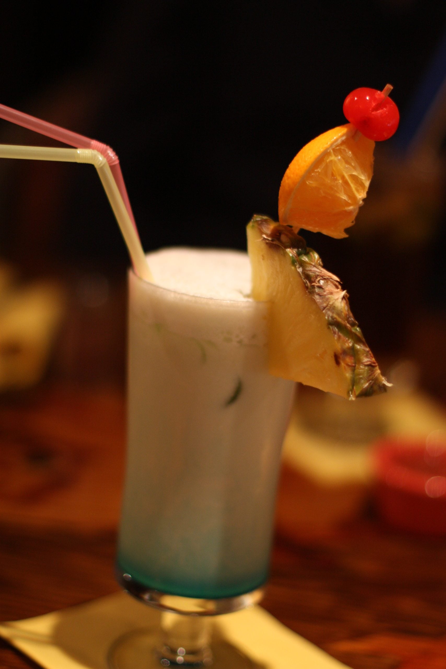 A tropical cocktail in a tall glass with a pineapple slice, orange wedge, and cherry garnish, topped with foam, and two straws.