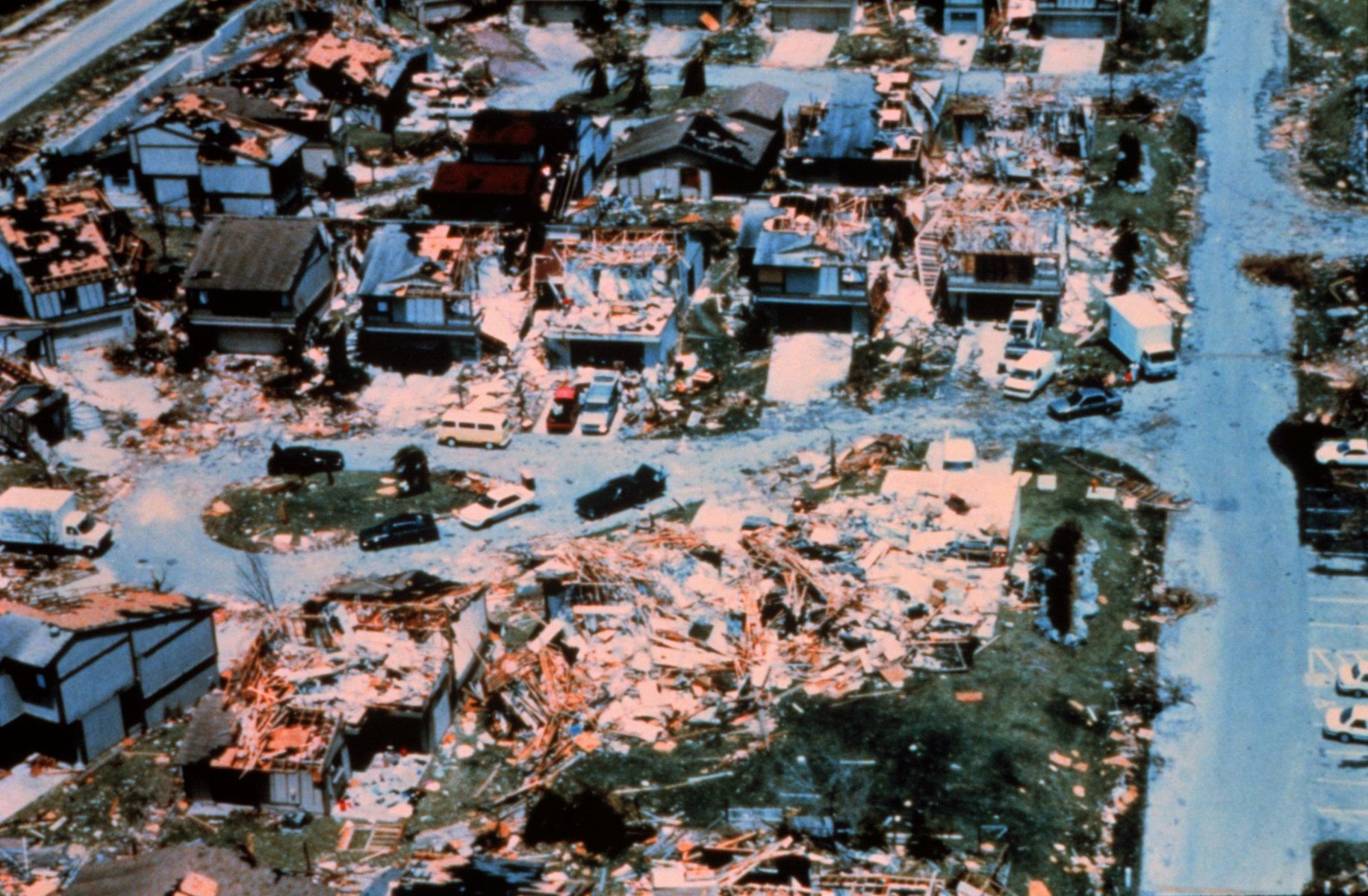 Aerial view of a residential neighborhood severely damaged by a natural disaster, with roofs missing, debris scattered, and homes destroyed. Vehicles are parked among the wreckage on cracked streets.