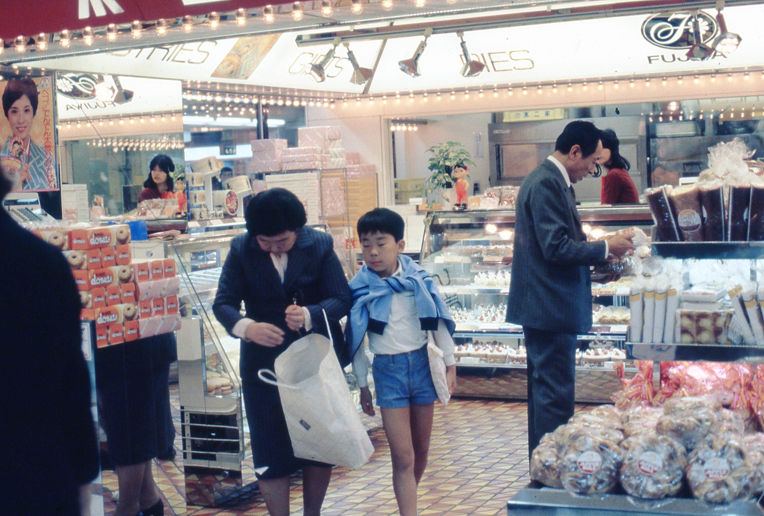 A woman and a boy shop in a brightly lit bakery, surrounded by pastries and bread, while a man in a suit looks at the display. Other shoppers and staff are in the background.