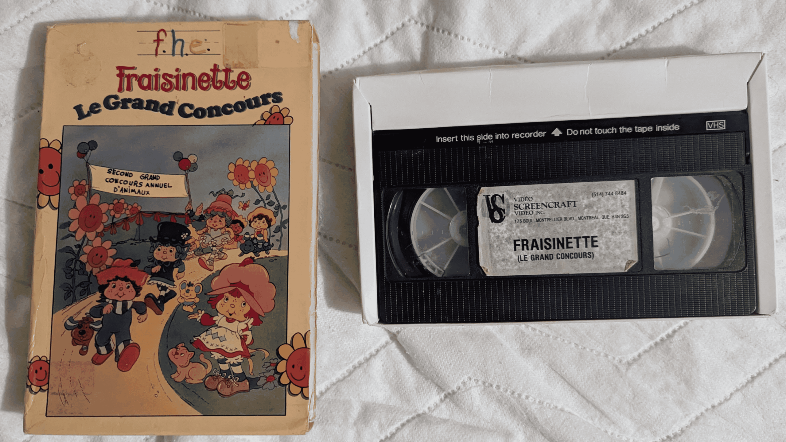 A vintage VHS tape and its illustrated box for “Fraisnette Le Grand Concours” rest on a white quilted surface. The cover shows cartoon characters among flowers, with French text and a handwritten note.
