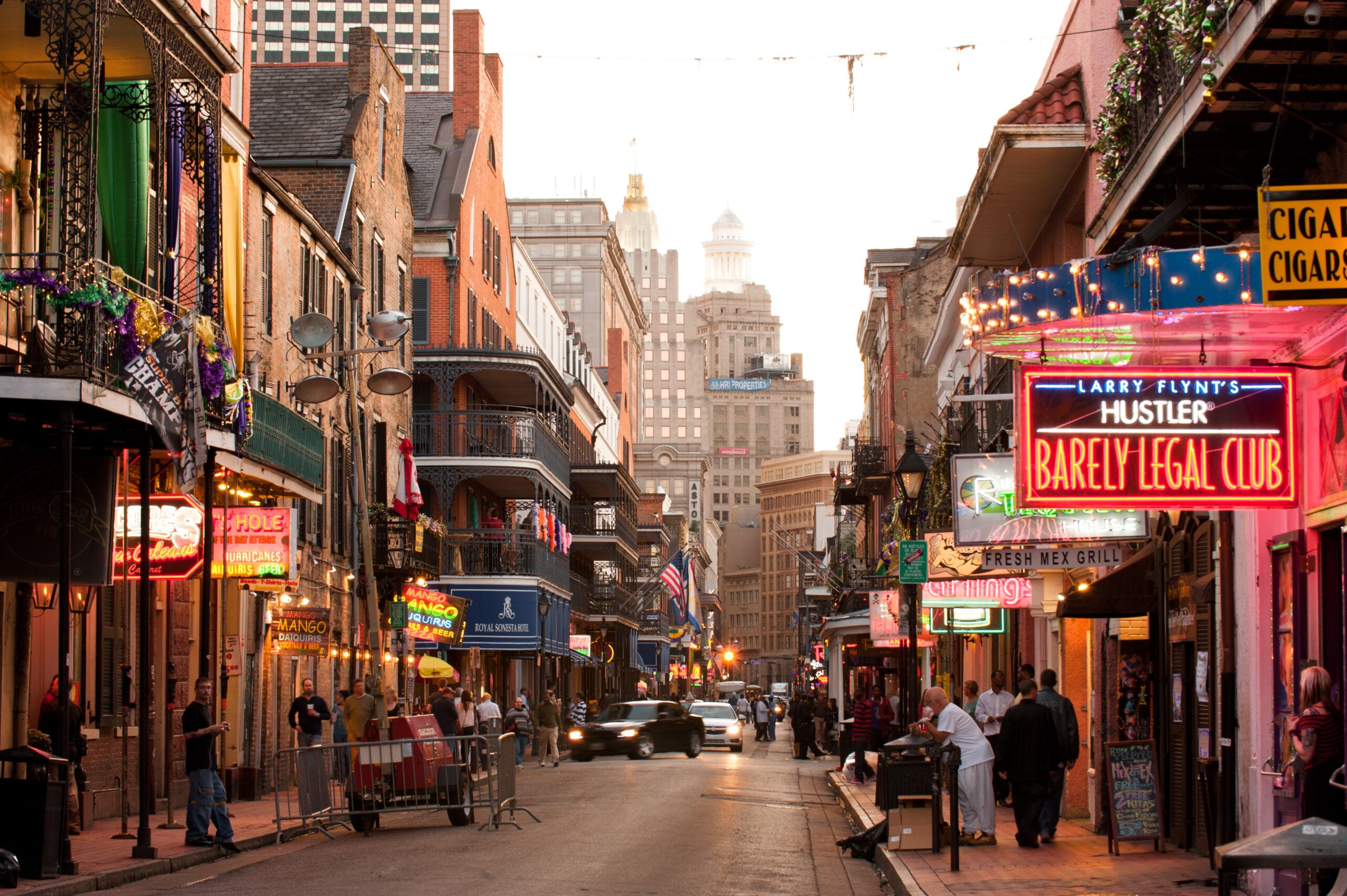 Bourbon Street in New Orleans at dusk, lined with colorful neon signs, bars, and people walking along the lively street. Skyscrapers are visible in the background.