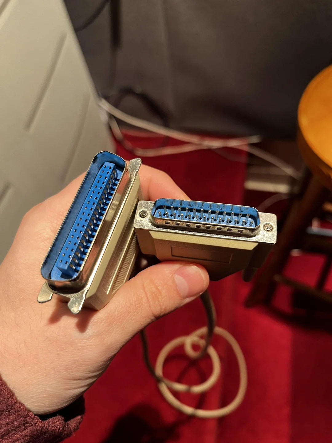 A hand holds two beige parallel port connectors with blue interiors, over a red carpet. One connector has more pins than the other. A wooden stool and cables are visible in the background.