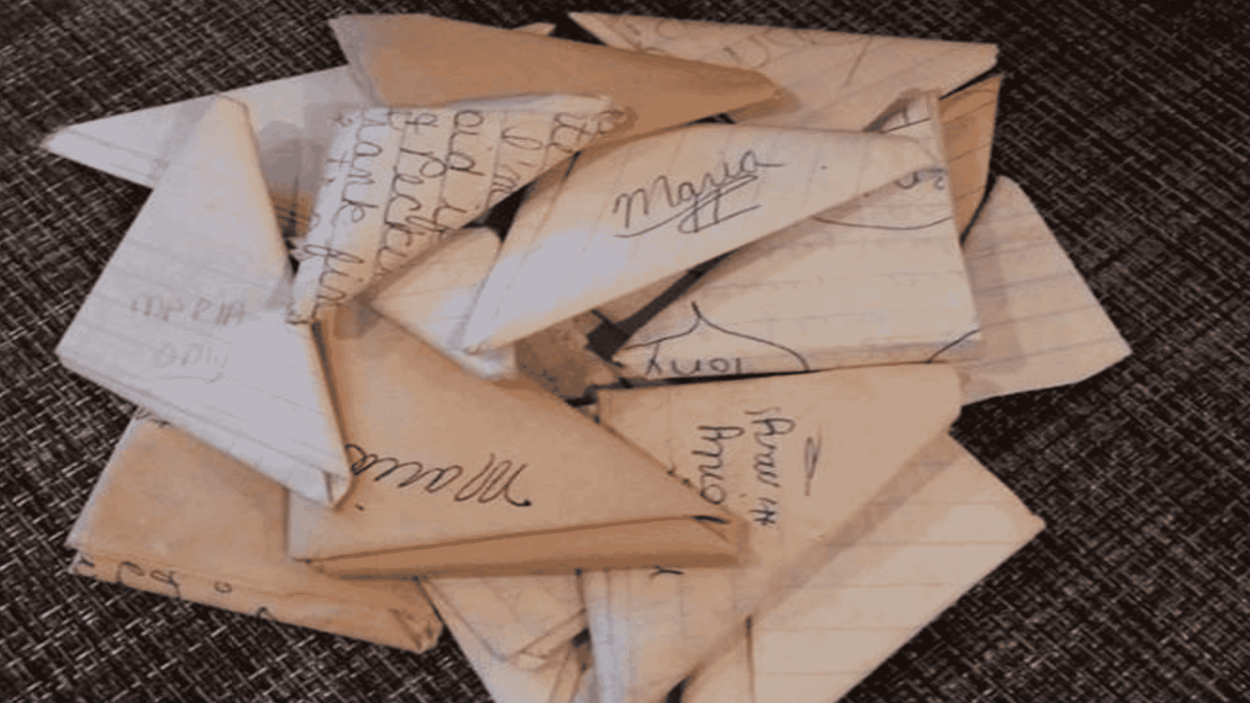 A pile of small, triangularly folded paper notes with handwritten text on them, some on lined and some on plain paper, resting on a dark textured surface.