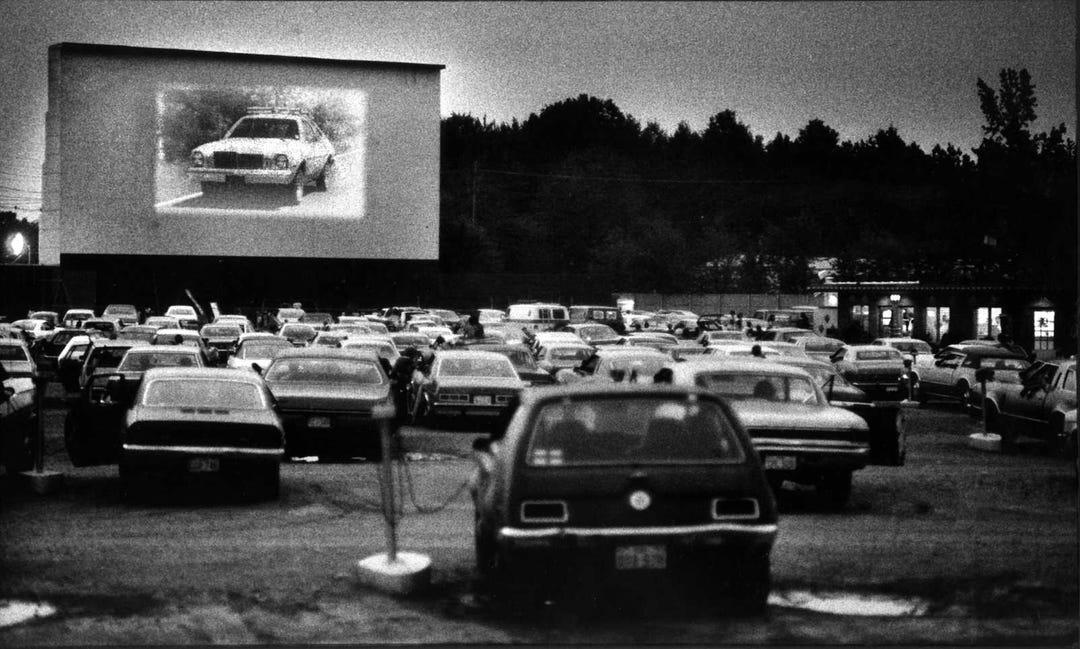 Black and white photo of a crowded drive-in theater at night, with cars parked facing a large outdoor movie screen showing a car chase scene. Trees and a concession stand are visible in the background.