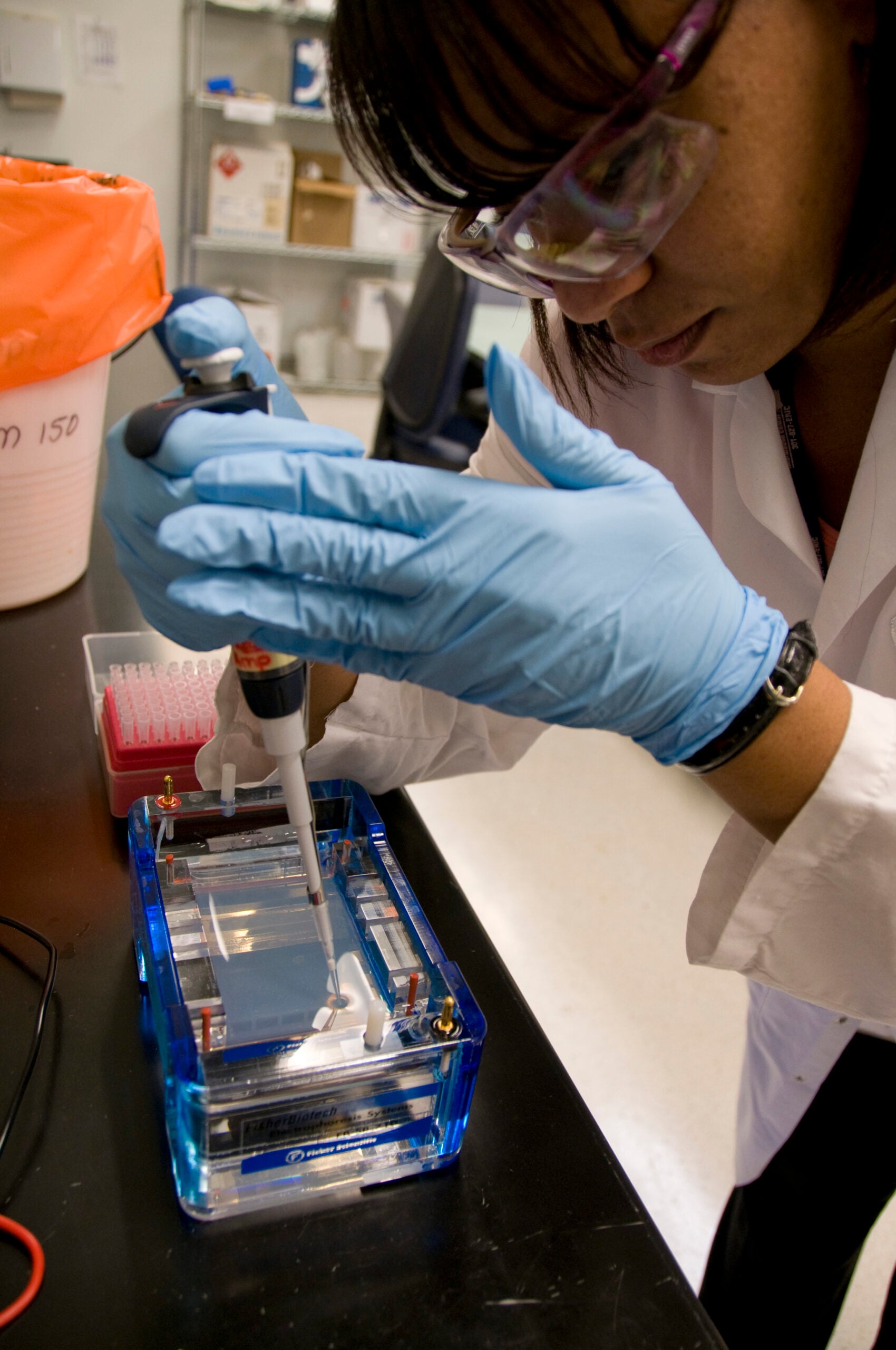 A scientist in safety glasses and blue gloves uses a pipette to load samples into a gel electrophoresis chamber in a laboratory setting.