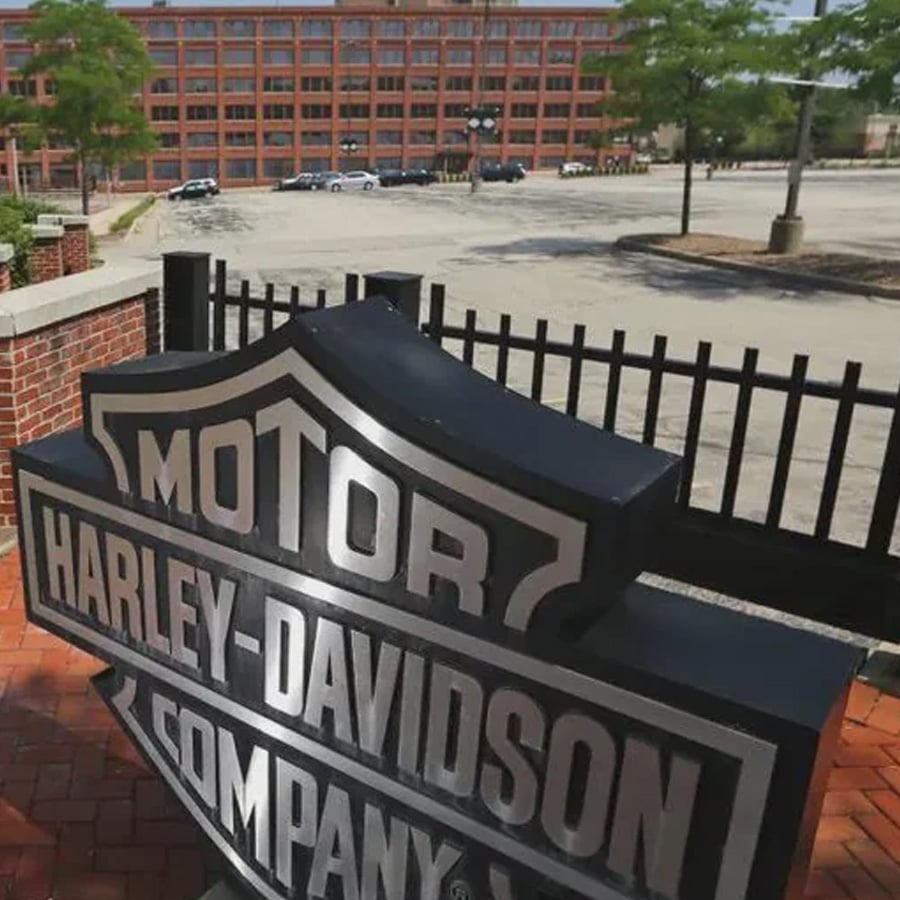 A large Harley-Davidson Motor Company sign stands in front of a fenced parking lot with a red brick industrial building and some trees visible in the background.