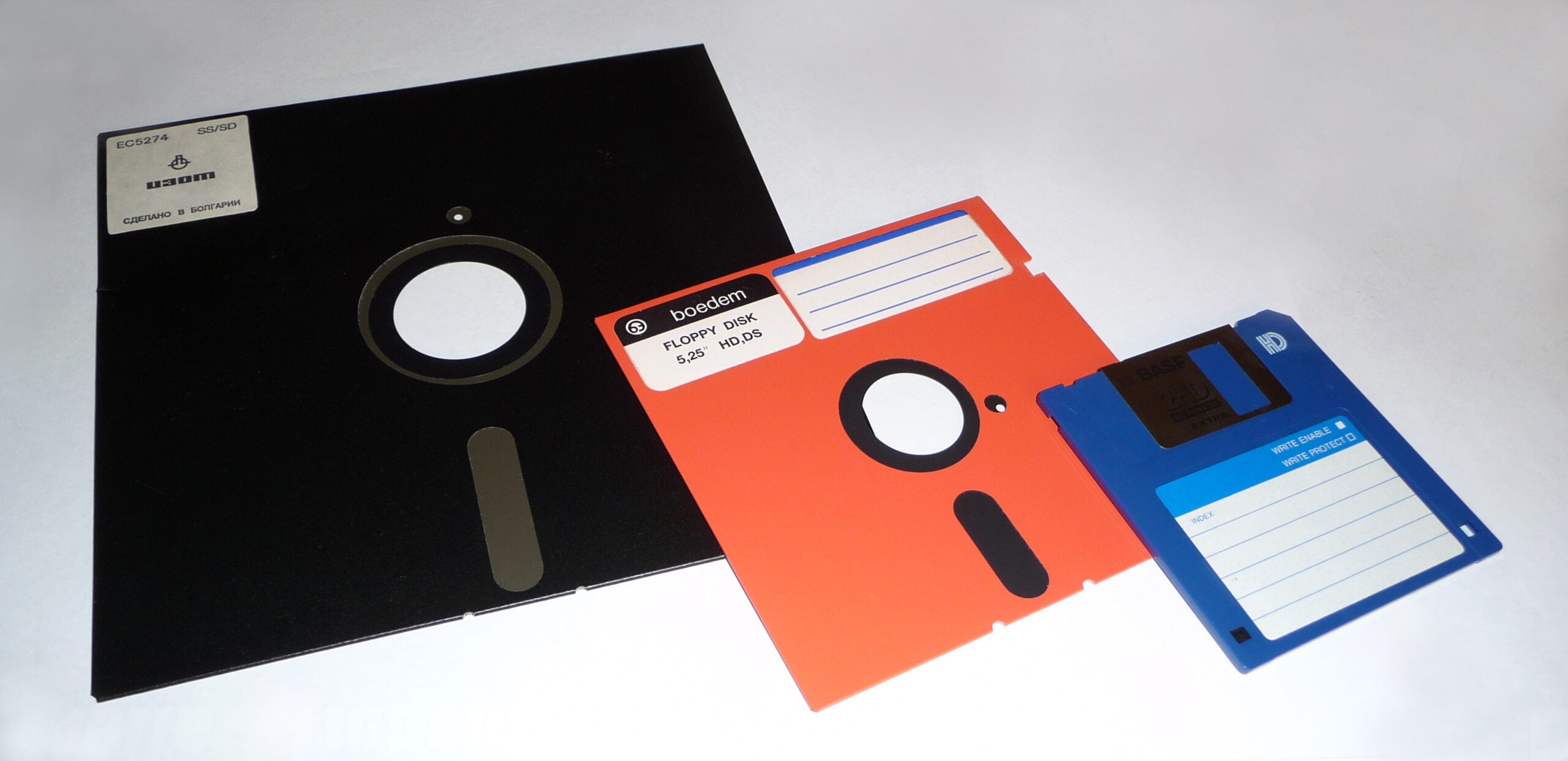 Three floppy disks of different sizes (8-inch black, 5.25-inch orange, and 3.5-inch blue) are arranged on a white surface, showing their labels and writable areas.