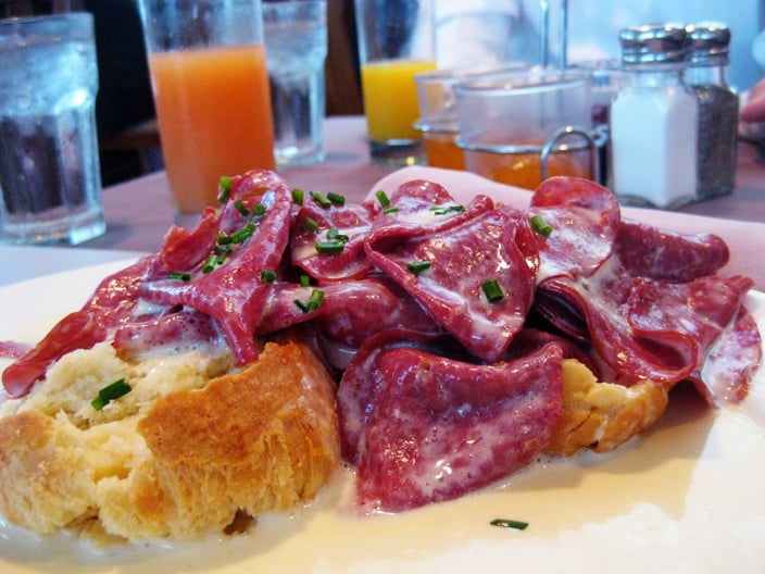 A biscuit topped with creamy sauce and slices of cured meat, garnished with chopped chives, sits on a table set with drinks and condiments.