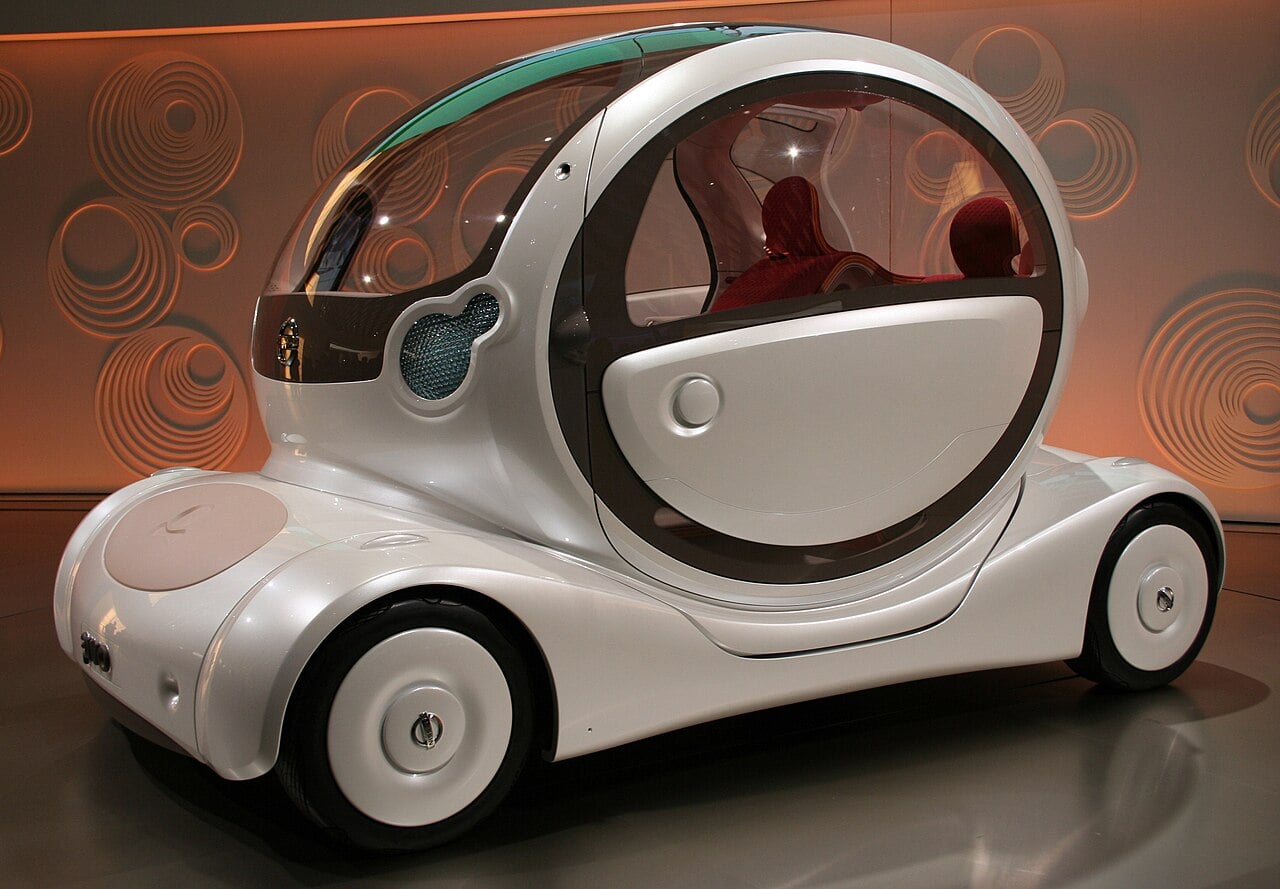 A futuristic, compact white concept car with a rounded, bubble-like shape and large windows is displayed indoors against a wall with circular patterns.