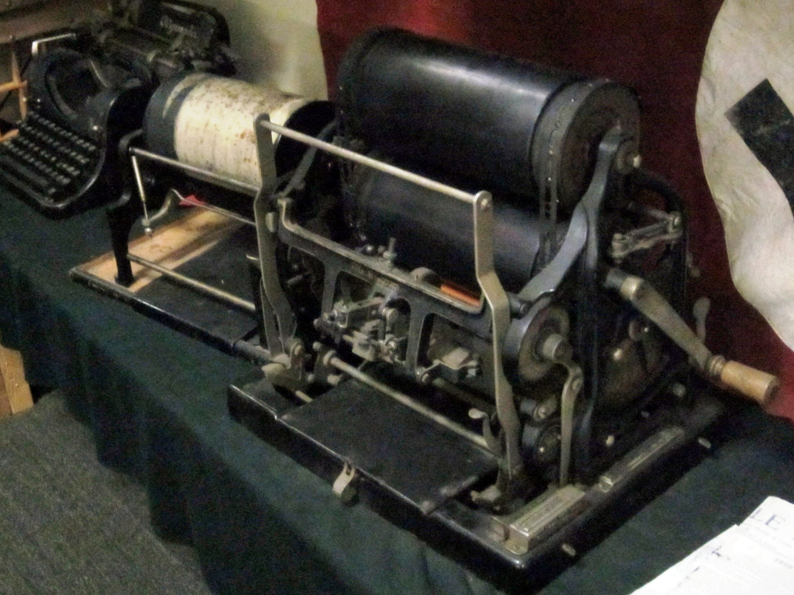 A vintage mimeograph machine with rollers and a hand crank sits on a table, next to an old typewriter.