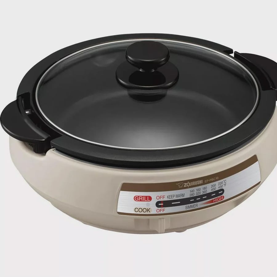 A round electric skillet with a glass lid, a white base, black handles, and a control panel labeled grill, cook, simmer, and keep warm settings, displayed against a plain background.