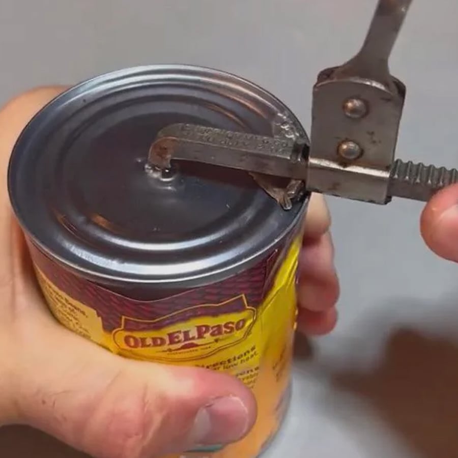 A hand uses a manual can opener to open a can of Old El Paso food. The opener is attached to the can’s lid, and the label is partially visible.
