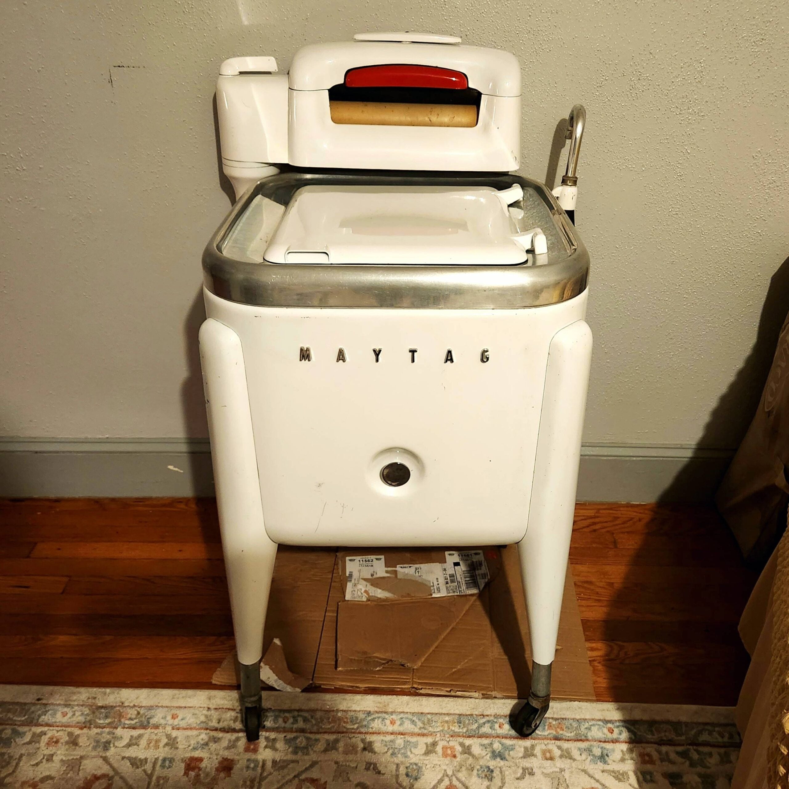 A vintage white Maytag wringer washing machine stands on wheels over a cardboard box, against a wall on a hardwood floor, next to a patterned rug.
