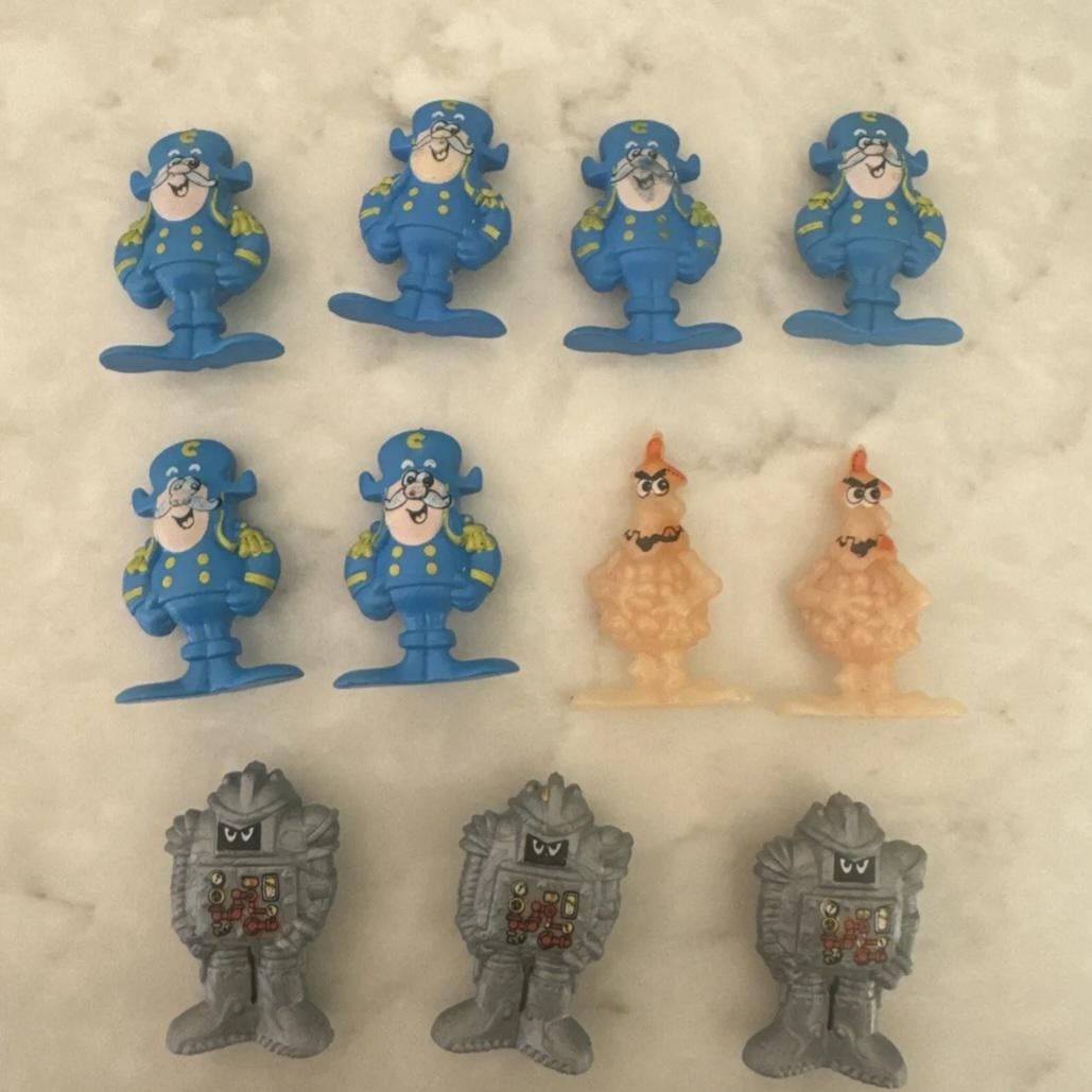 Nine colorful toy figures are arranged on a white surface: six blue captains, two tan chickens, and three gray robots, each with distinctive details and expressions.