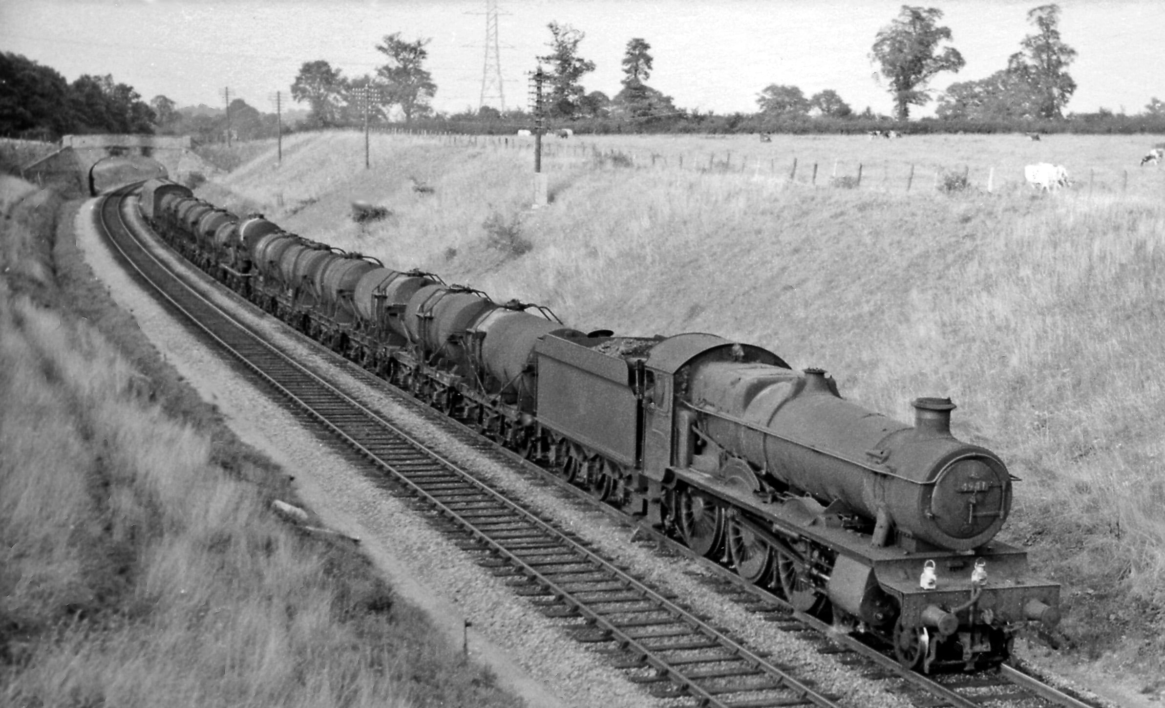 A vintage steam locomotive pulls a long train of cylindrical tank cars along a curved railway track through grassy countryside, with trees and utility poles in the background.