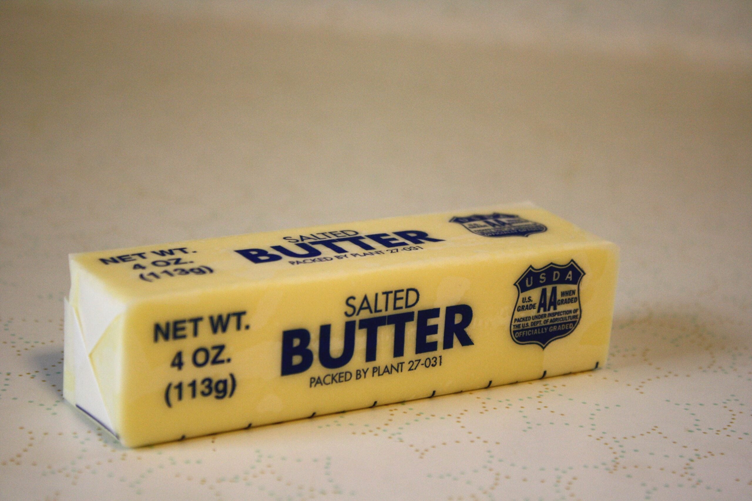 A stick of salted butter in its wrapper sits on a light patterned surface. The label reads “Salted Butter,” “Net Wt. 4 oz (113g),” and shows a USDA AA quality shield.