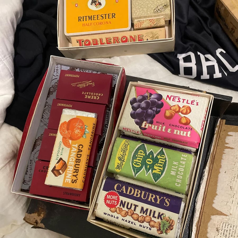Two open boxes filled with vintage chocolate and candy bars, including Cadbury’s, Nestlé, Toblerone, and Ritmeester, rest on a bed near dark clothing and books.