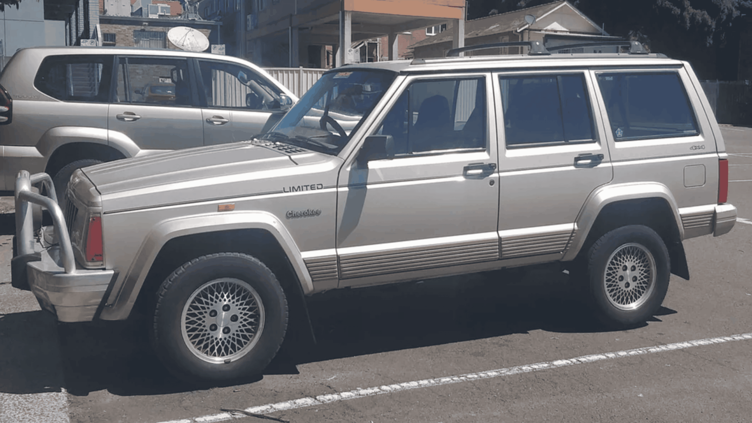 A beige Jeep Cherokee Limited SUV is parked in a lot on a sunny day, with another vehicle parked nearby and buildings visible in the background.