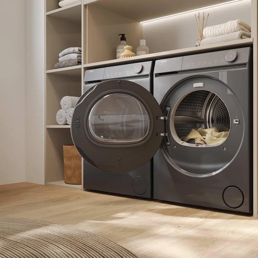 Two modern front-loading washing machines sit side by side in a laundry room. One machine door is open with laundry inside. Shelves above hold towels, bottles, and decor items. The room has wood flooring.
