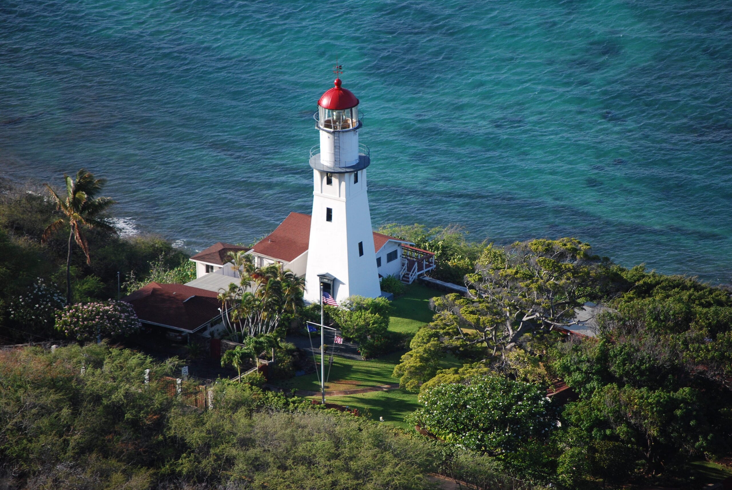 A white lighthouse with a red roof stands by the blue ocean, surrounded by green trees and small buildings, viewed from above.