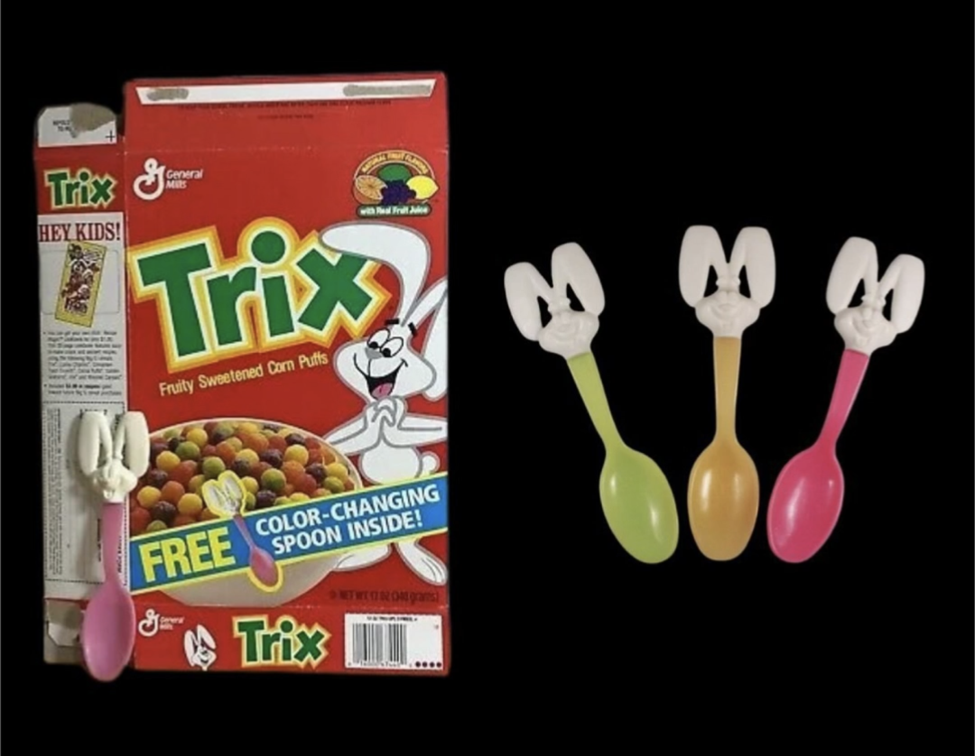 A Trix cereal box with colorful cereal, plus three color-changing spoons—yellow, green, and pink—each topped with a bunny head, displayed against a black background.