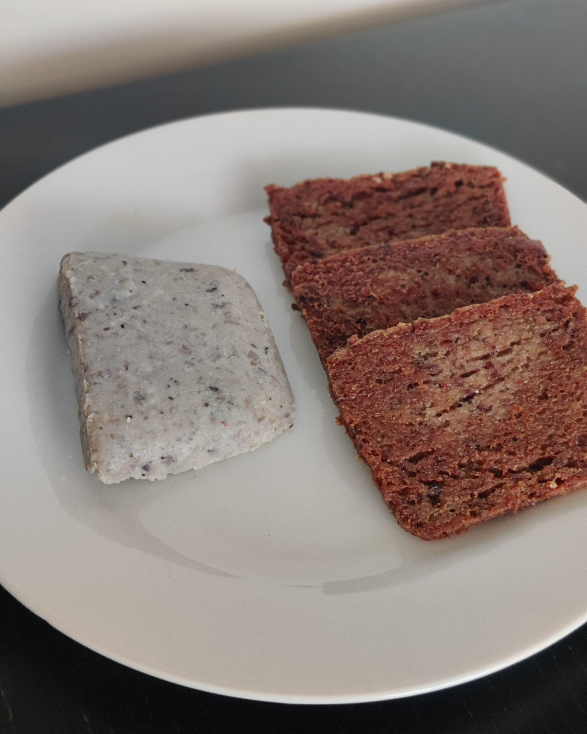 A white plate with three slices of brown bread and a rectangular piece of light gray food speckled with dark spots, set on a dark surface.