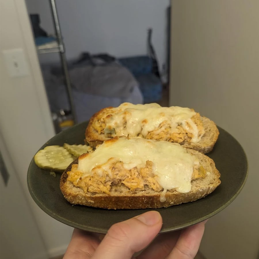A hand holds a plate with two slices of bread topped with shredded chicken and melted cheese, served with two pickle slices on the side.