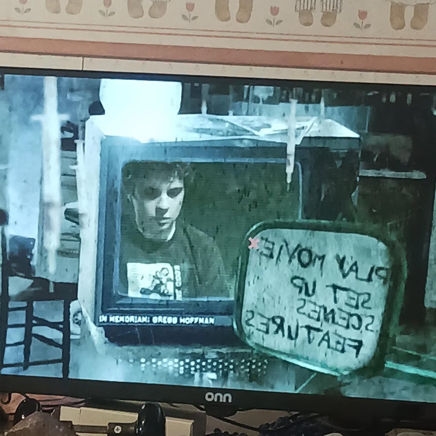 A person’s face appears on an old TV monitor. A small whiteboard with the message “PLAY MOVIE, SET UP SCENES, FEATURES” is written backward. The screen reads “IN MEMORIAM: GRACE HOFFMAN.”