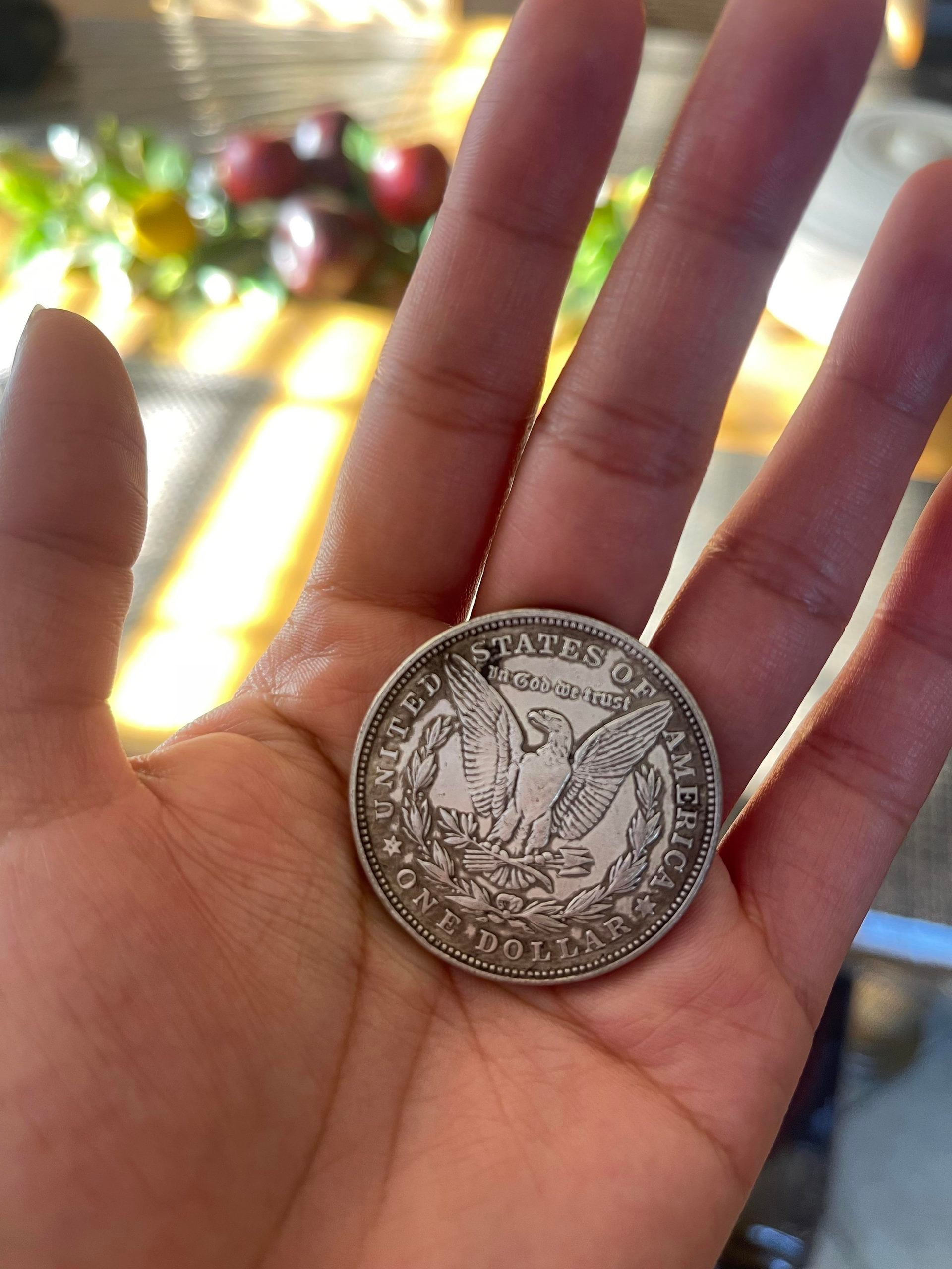 A hand holds a silver U.S. dollar coin featuring an eagle with spread wings and the words “United States of America” and “One Dollar” visible.