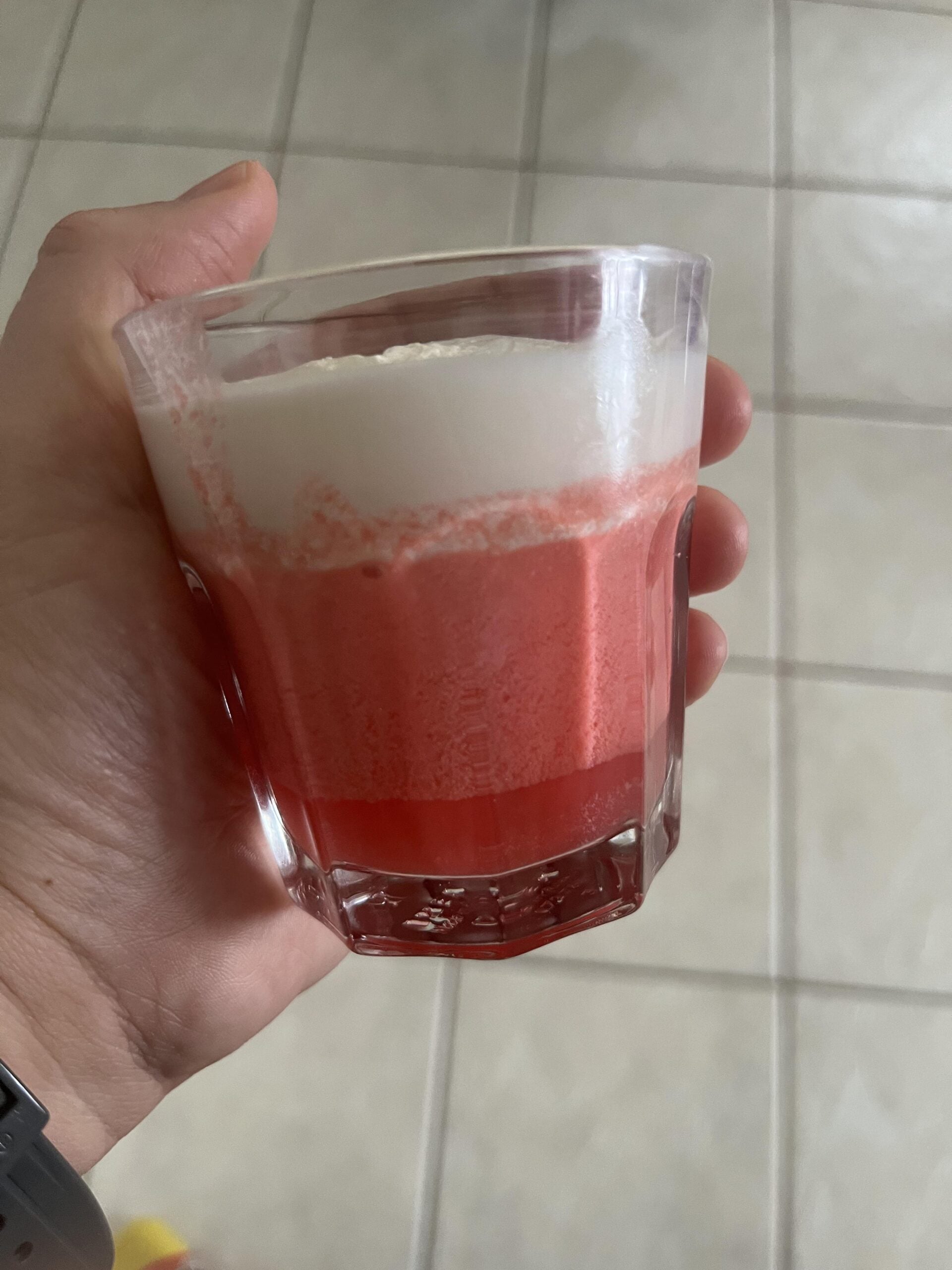 A hand holds a glass with a layered drink: a thick, frothy white layer on top of a vibrant pink layer. The background shows a light-colored tiled floor.