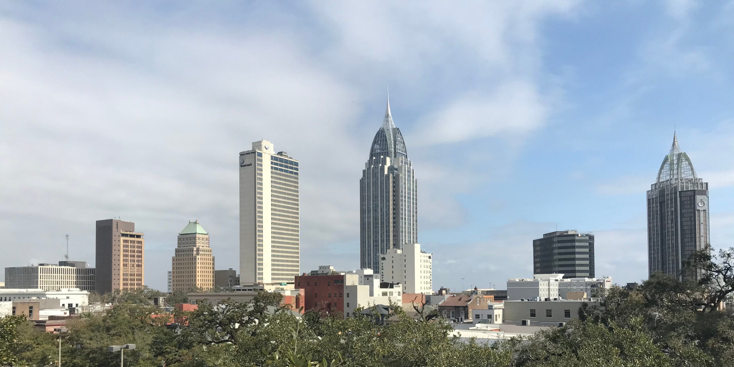 Skyline of a city with several tall buildings, including two with pointed tops, against a partly cloudy blue sky. There are shorter buildings and green trees in the foreground.