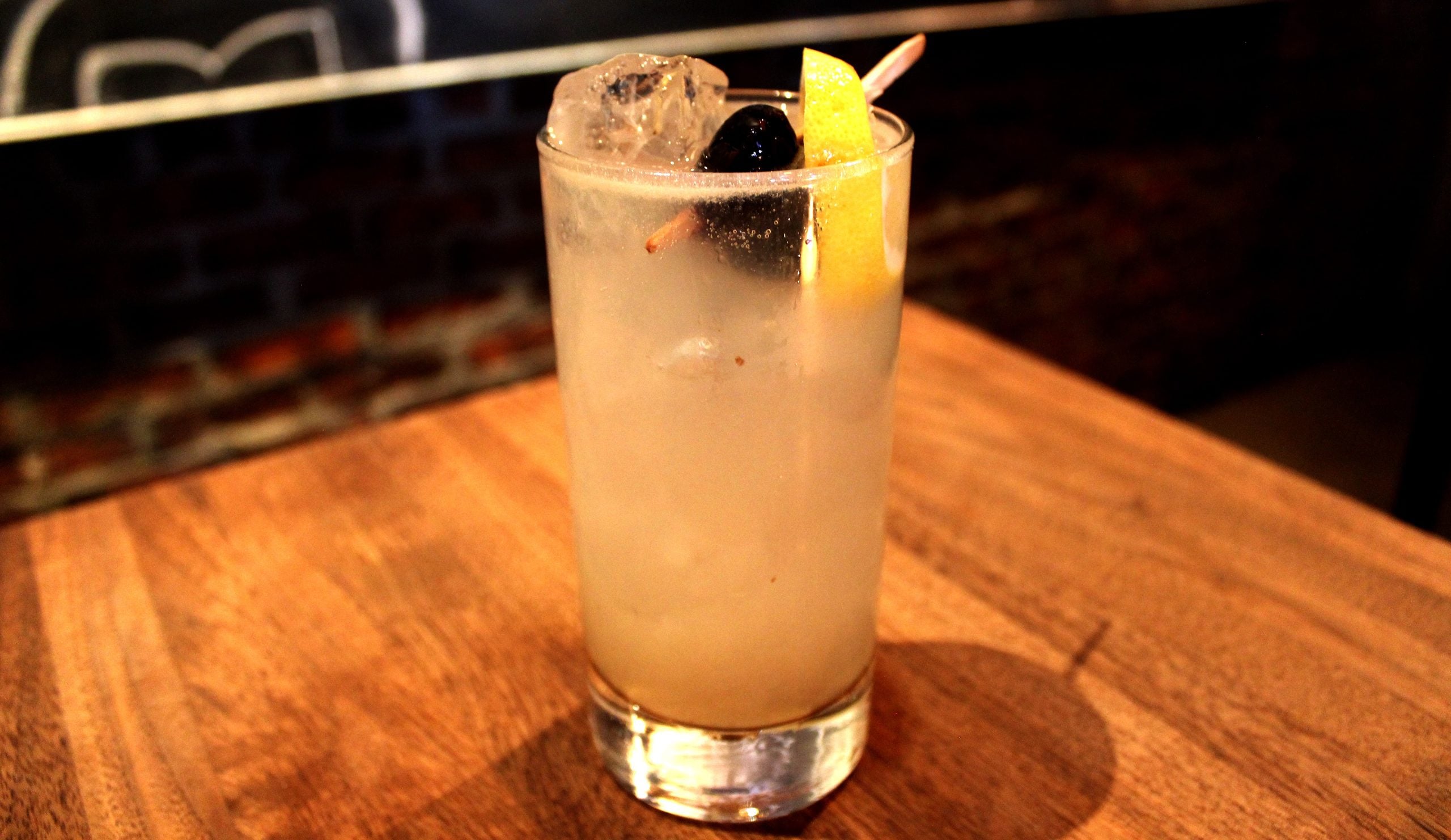 A tall glass with a light, cloudy drink, topped with ice, a lemon peel, and a cherry on a toothpick, sits on a wooden table.