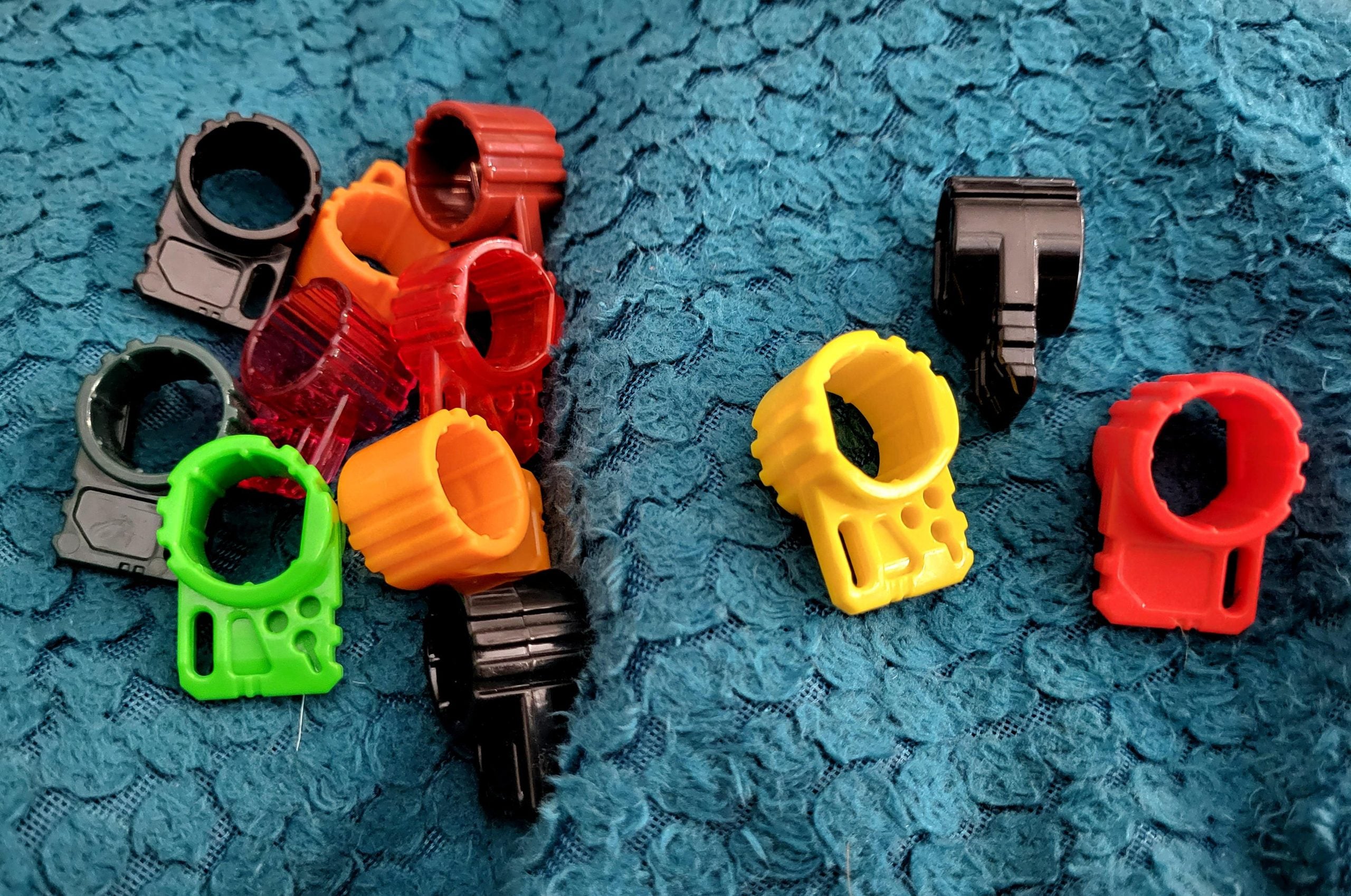 Colorful plastic bread bag clips, including black, red, green, yellow, and orange, are scattered on a textured blue fabric surface. Two clips, one yellow and one red, are separated from the main group.