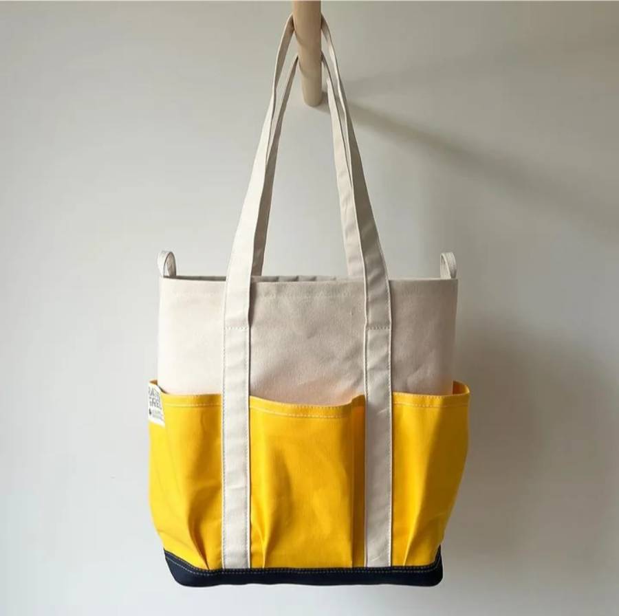A canvas tote bag with cream handles and upper section, yellow pockets, and a dark blue base, hanging on a wooden hook against a plain light background.