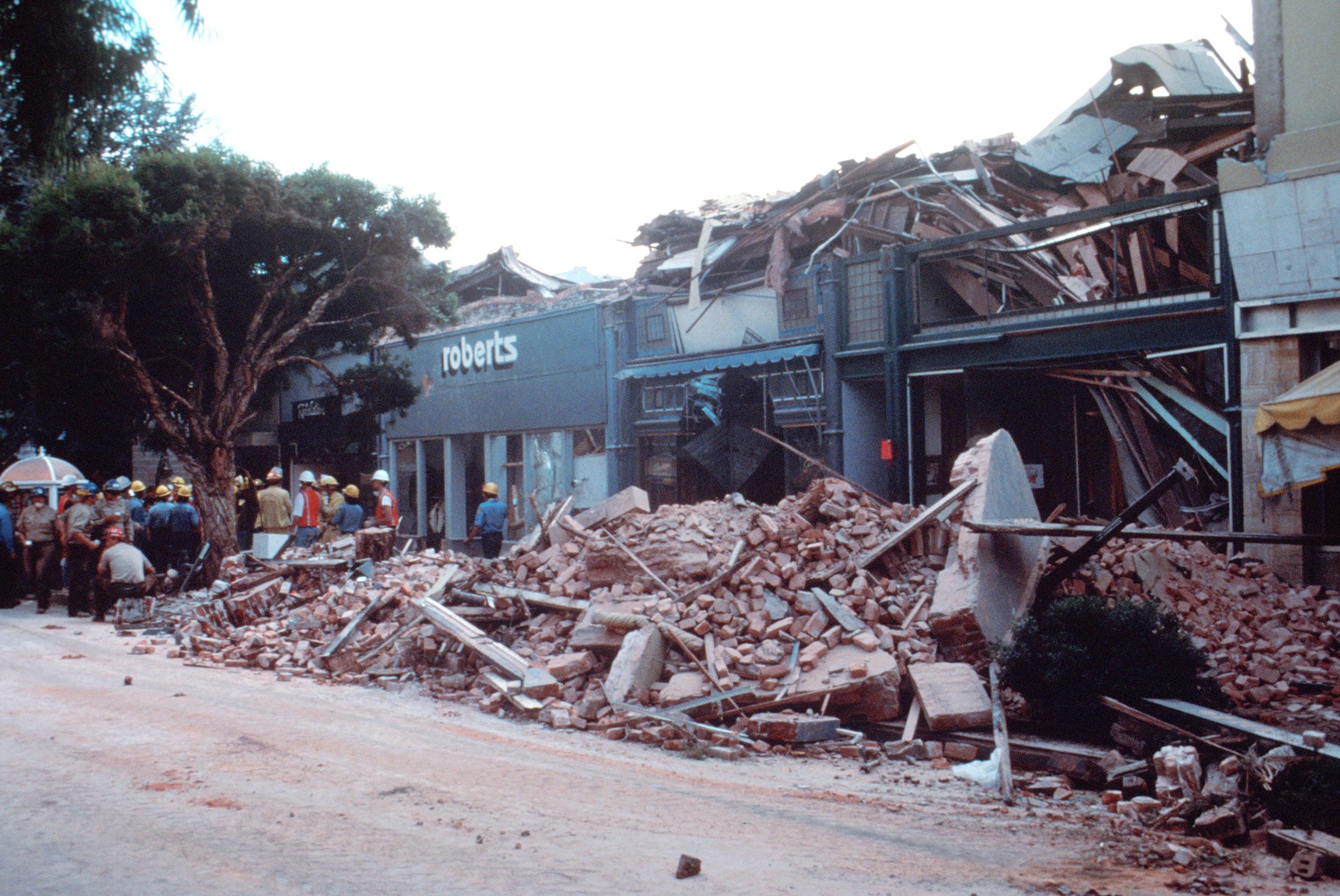 A group of rescue workers stand near the rubble of a collapsed building on a city street, surrounded by debris after a disaster.