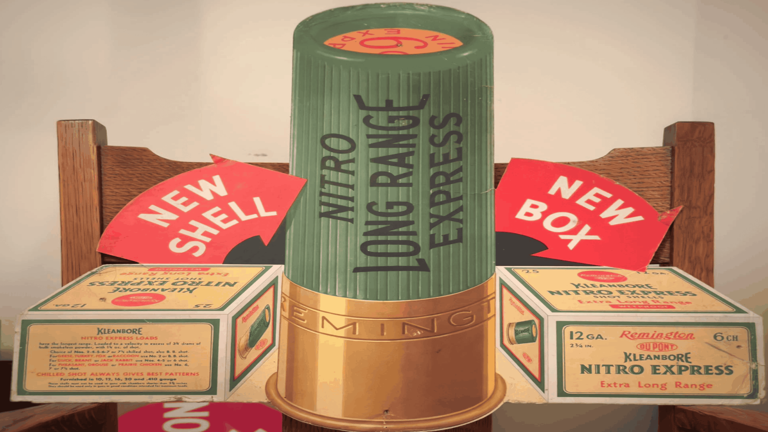 A vintage Remington Nitro Express shotgun shell display with a large shell replica, two red “NEW SHELL” and “NEW BOX” signs, and several boxes of 12-gauge Remington Kleanbore shells on a wooden chair.