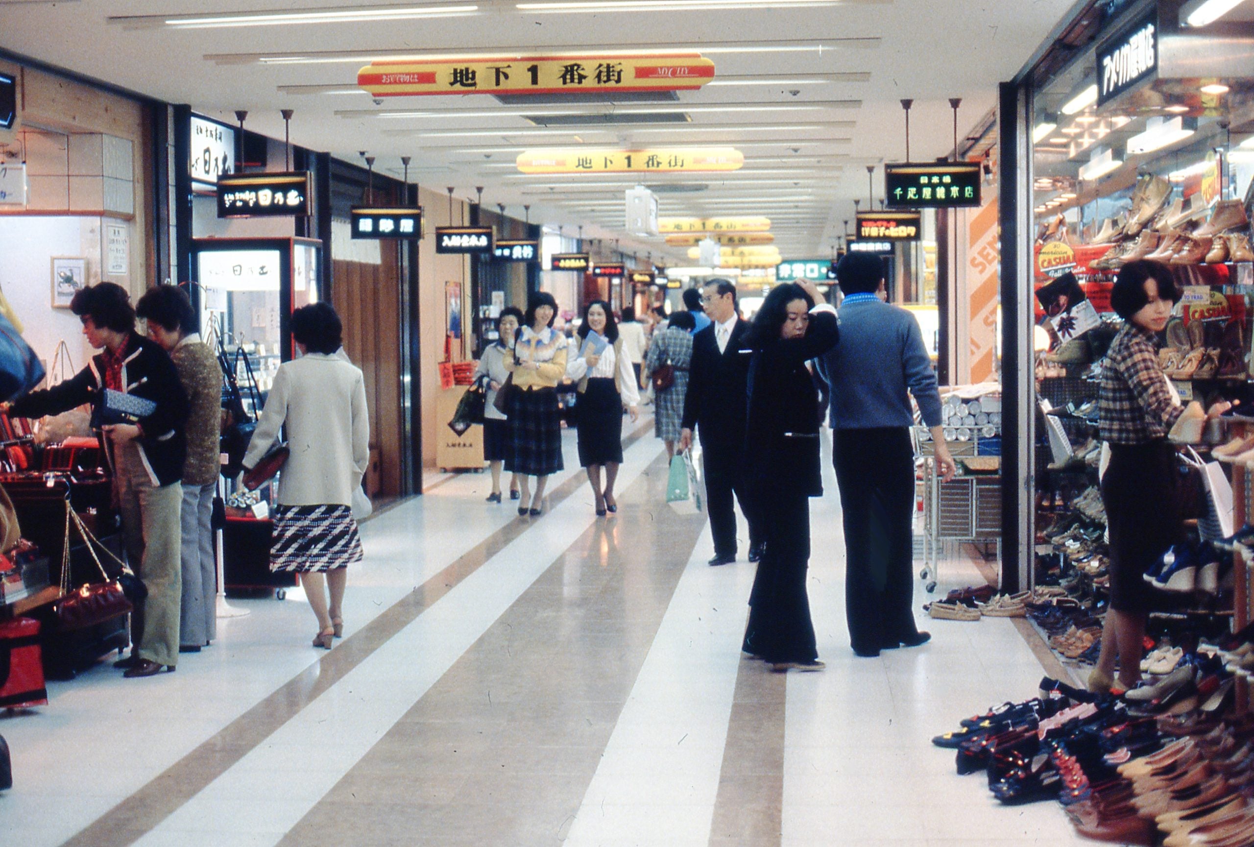 Shoppers walk through a brightly lit indoor mall lined with shoe and clothing stores, with signs in Japanese hanging from the ceiling and people browsing merchandise.