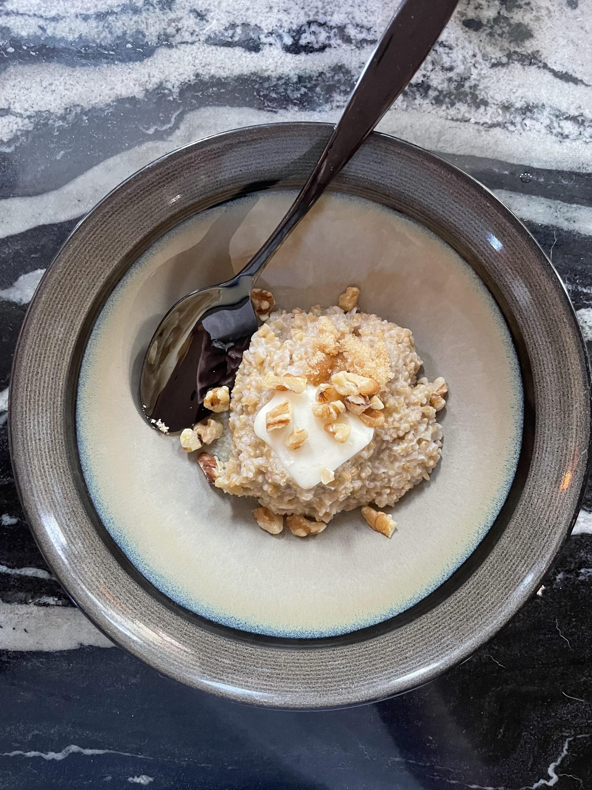 A bowl of oatmeal topped with a pat of butter and chopped walnuts, with a spoon resting in the bowl, placed on a black and white marble surface.