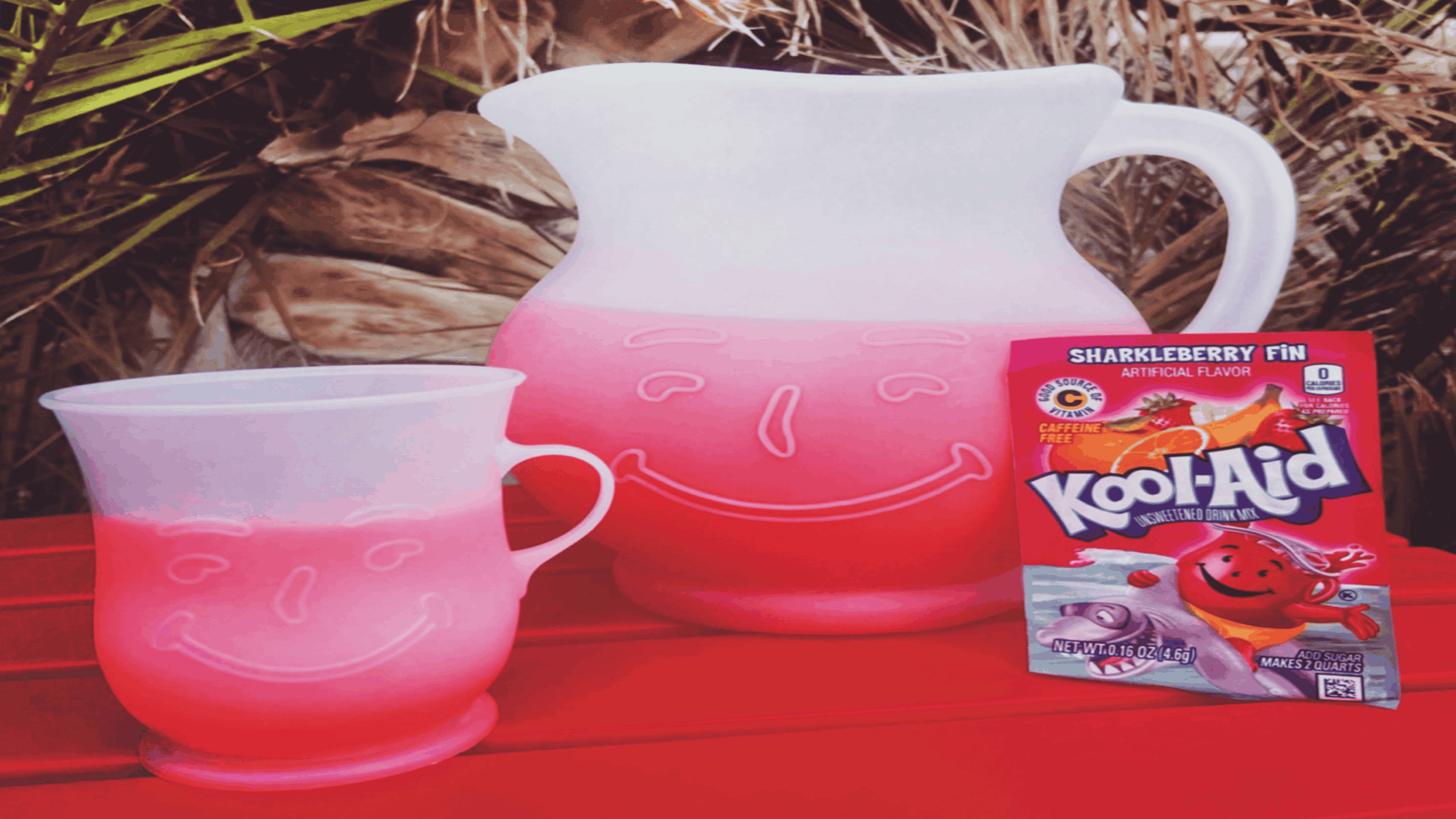 A pink Kool-Aid pitcher and cup with smiling faces sit on a red table beside a packet of Sharkleberry Fin Kool-Aid, with plants in the background.
