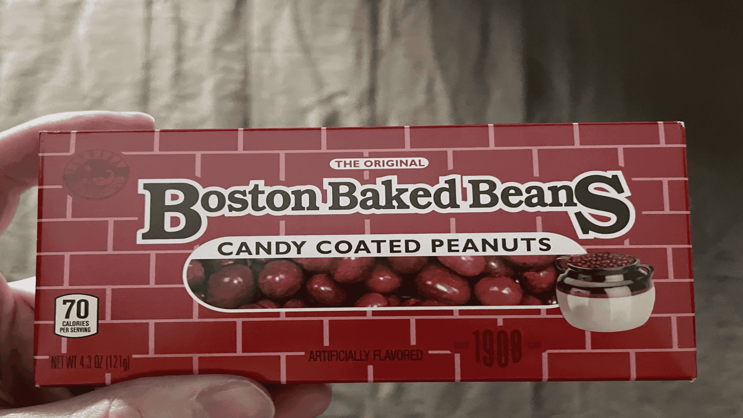A hand holds a red box of Boston Baked Beans candy-coated peanuts. The box has a brick design, an image of the candy, and states “70 calories per serving.”