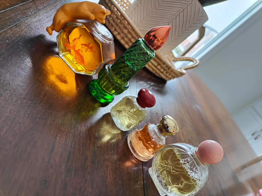 Five ornate perfume bottles, some with colorful caps, are arranged on a wooden surface near a woven basket and a white vase in a sunlit room.