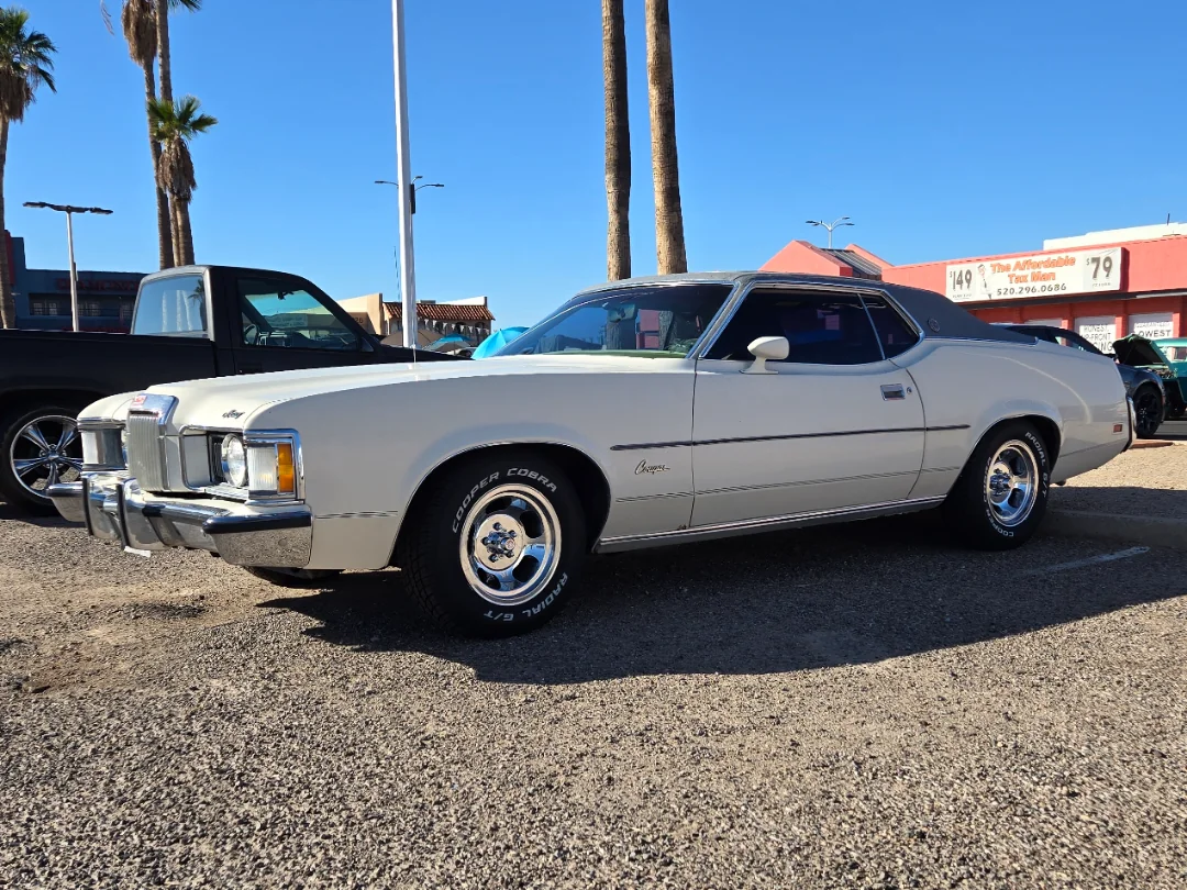 A white classic Mercury Cougar car is parked in a lot on a sunny day, with palm trees and other vehicles in the background.