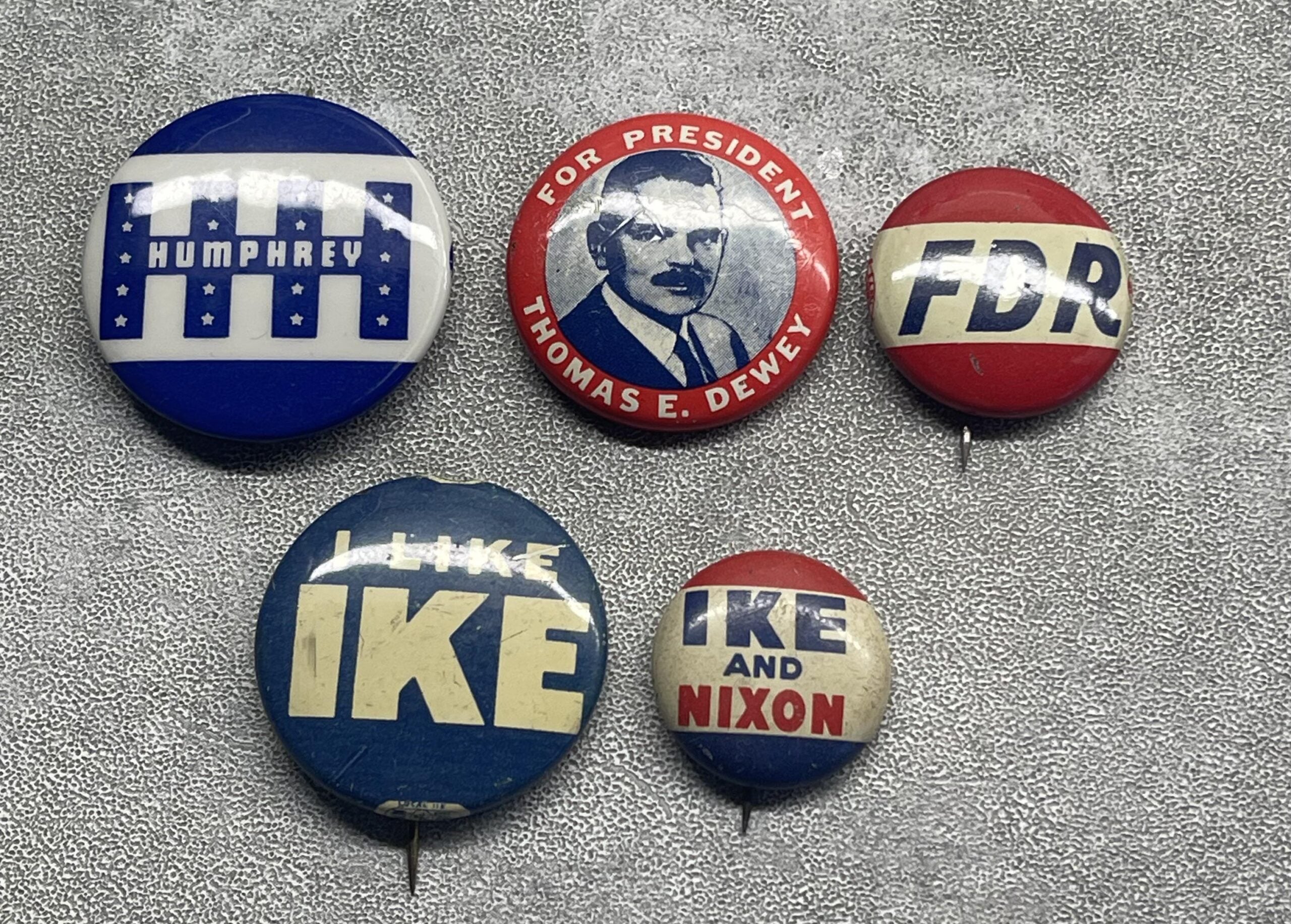 Five vintage U.S. presidential campaign buttons for Humphrey, Dewey, FDR, Eisenhower (“I Like Ike”), and “Ike and Nixon” on a textured gray surface.