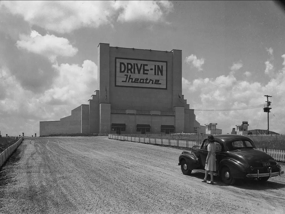 A woman stands by a vintage car on a gravel road in front of a large, retro drive-in theater building under a partly cloudy sky.