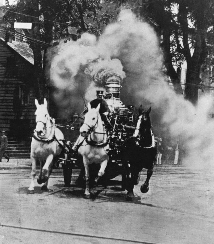 Three horses pull a steam-powered fire engine down a street, with a large plume of smoke billowing behind them. Trees and buildings line the background. The scene is from an earlier era.